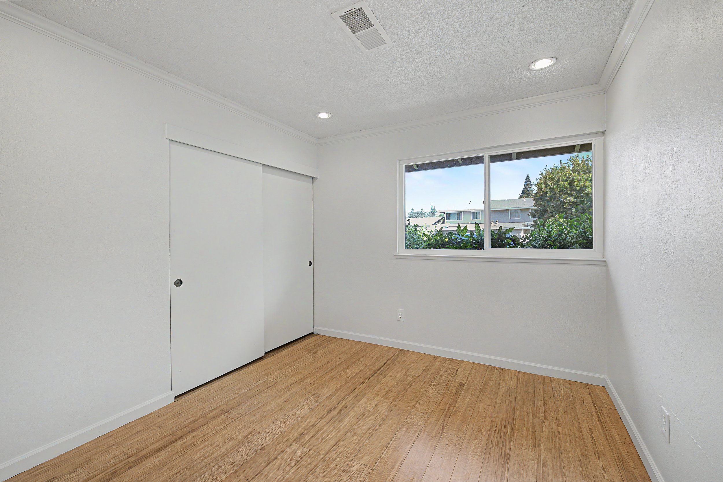 Empty room with light-colored walls, a window showing trees and buildings outside, a white closet door, crown molding, and wood flooring.
