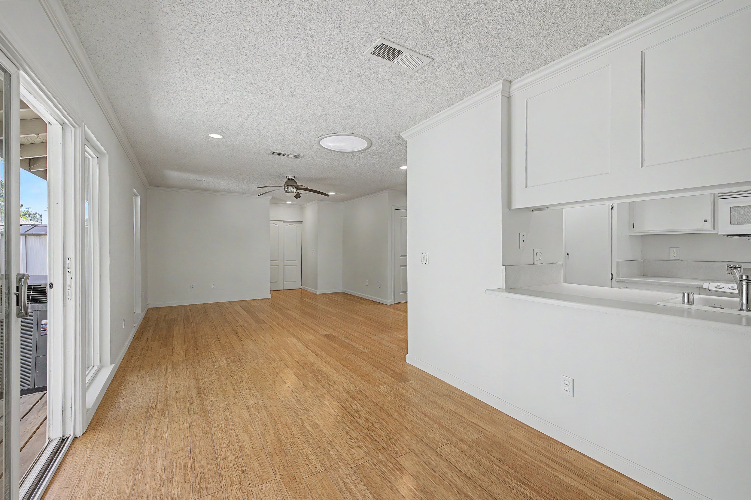 Empty open-concept living space with white walls, wood flooring, a ceiling fan, and a kitchen area with white cabinets and counters. A sliding glass door leads to an outdoor area.