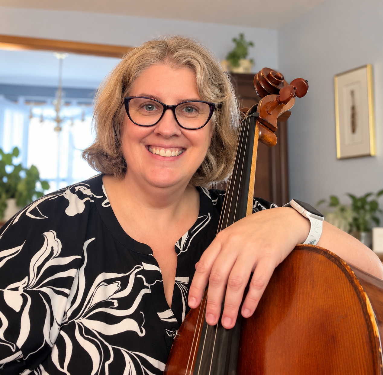A woman with glasses smiling while holding a cello in a bright, home interior.
