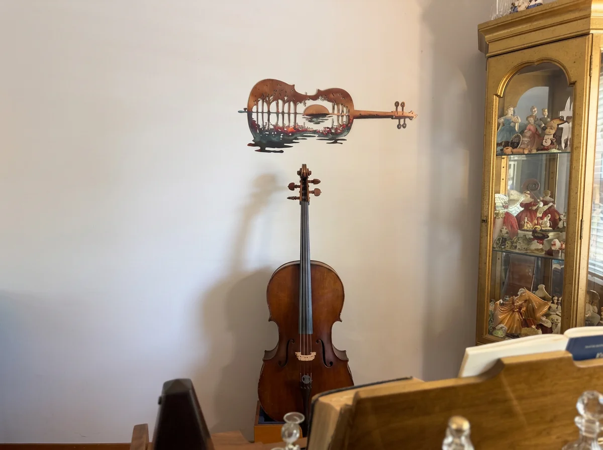 A wooden cello standing upright on the floor, with a decorative wall art piece shaped like a guitar hanging above it. To the right, there is a glass display cabinet with figurines and collectibles, and part of a wooden piece of furniture with sheets of paper or music.