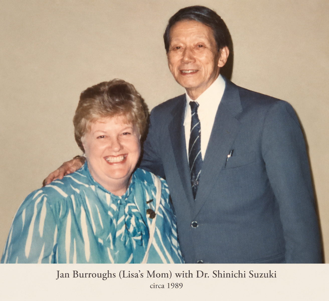 A woman and a man smiling, standing close together. The woman is wearing a blue and white striped blouse and a brooch, and the man is wearing a blue suit, white shirt, and striped tie. They are indoors, and the caption indicates they are Jan Burroughs (Lisa's mom) and Dr. Shinichi Suzuki, circa 1989.