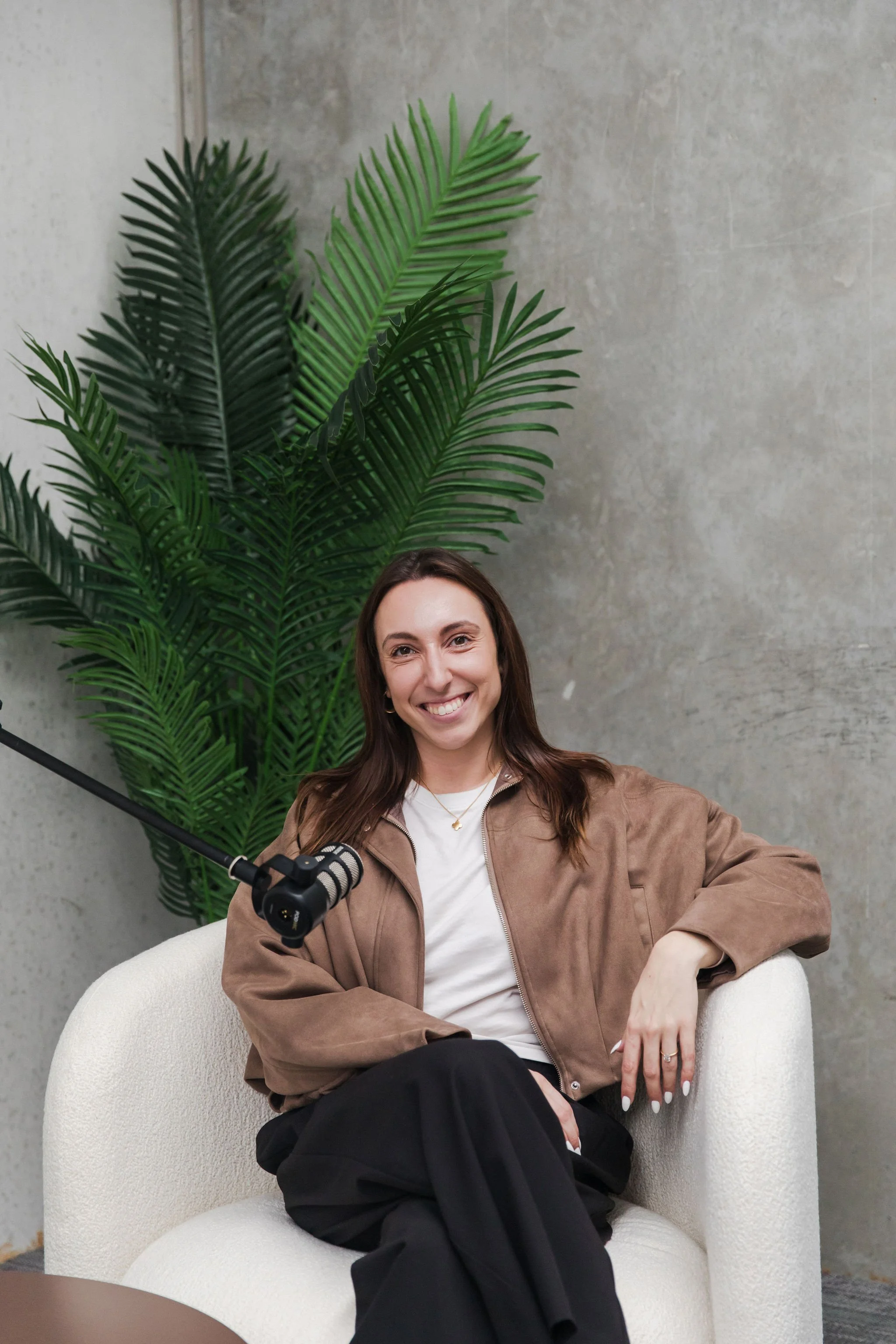 A smiling woman with shoulder-length brown hair, wearing a light brown jacket, white shirt, and black pants, sitting on a cream-colored armchair in front of a green potted plant and textured gray wall, holding a microphone.