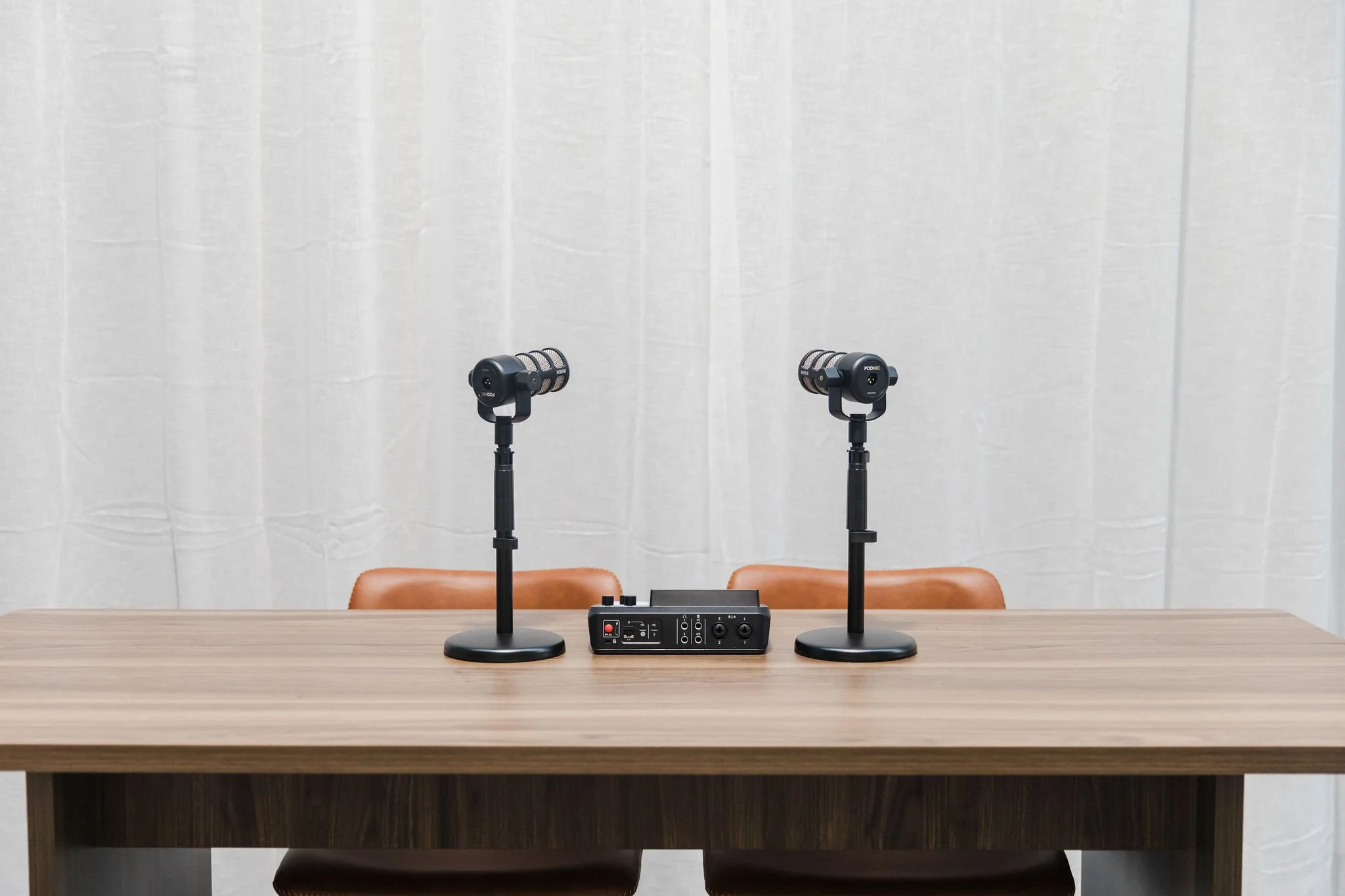 Two microphones on stands set up on a wooden table, with an audio interface device in front, against a white curtain backdrop.