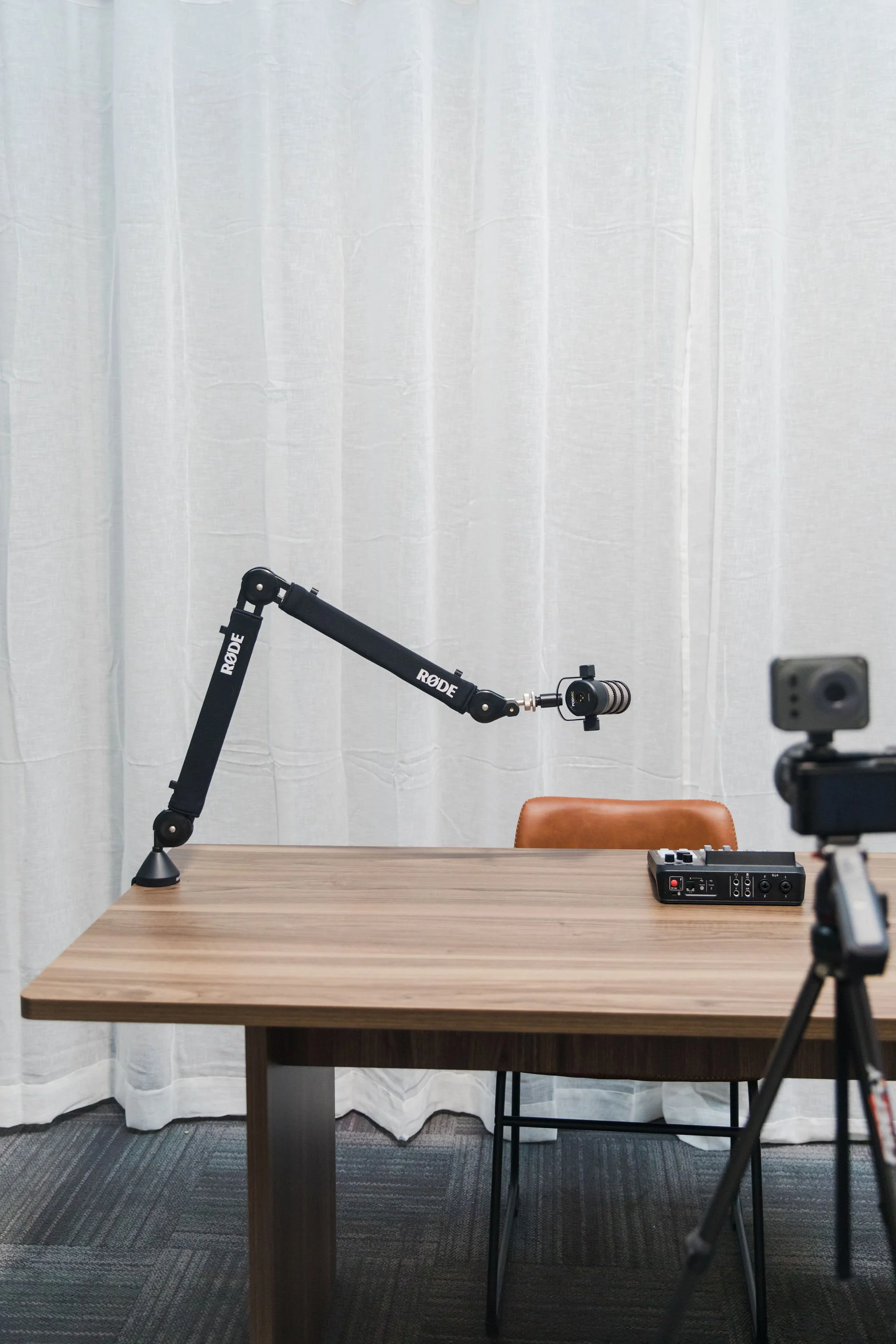 A wooden table with a brown chair, a black audio mixer, and a microphone arm with a microphone, against a white curtain backdrop, in a recording or podcast studio.