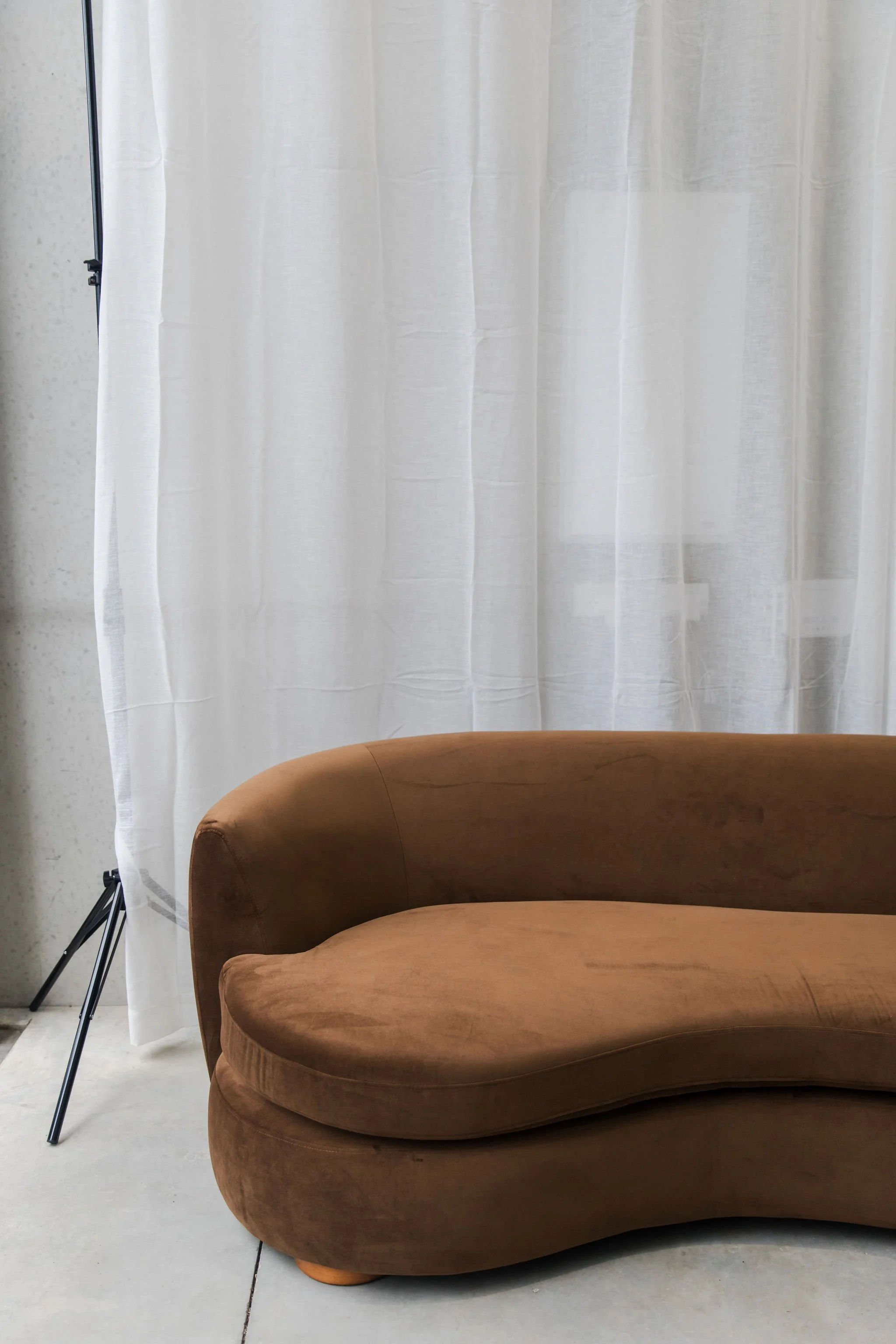 Brown velvet sofa in front of a white curtain with a metal tripod on the left side.