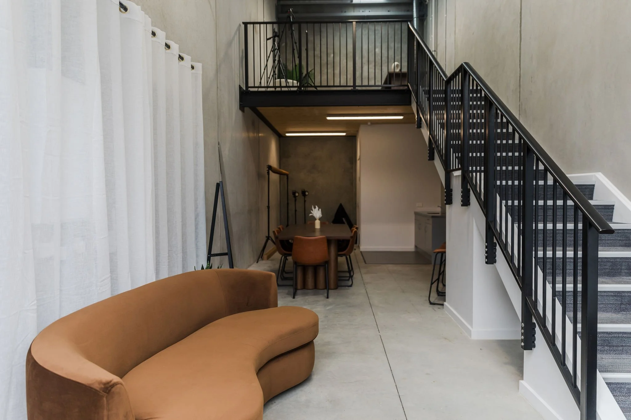 Modern interior space with a brown curved sofa in the foreground, white curtains on the left, a dining table with four orange chairs and a white vase with flowers nearby in the background, a staircase with black railing, and a small balcony with a tripod and plant on it.