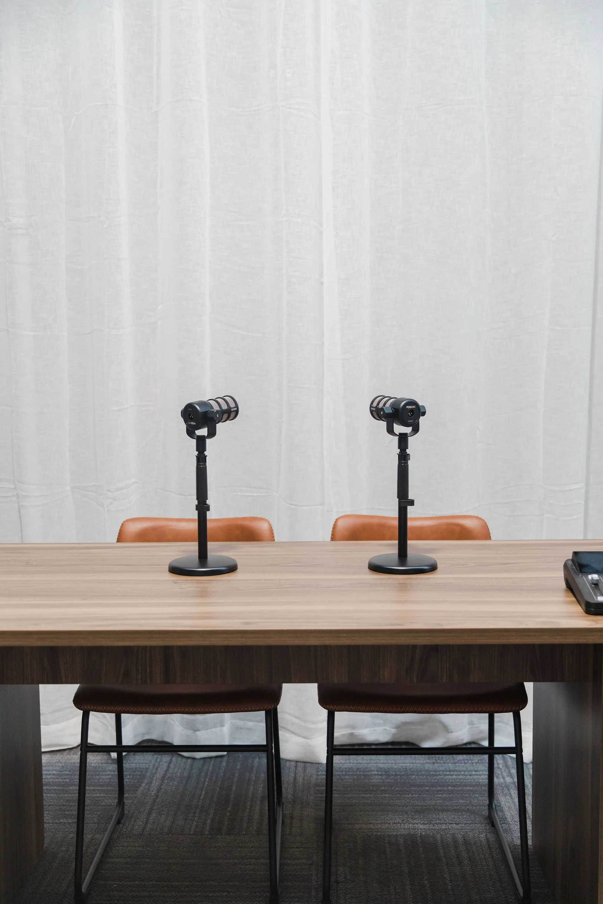 A conference room setup with two microphones on stands on a wooden table, two brown chairs, white curtains in the background, and a conference phone on the right side of the table.