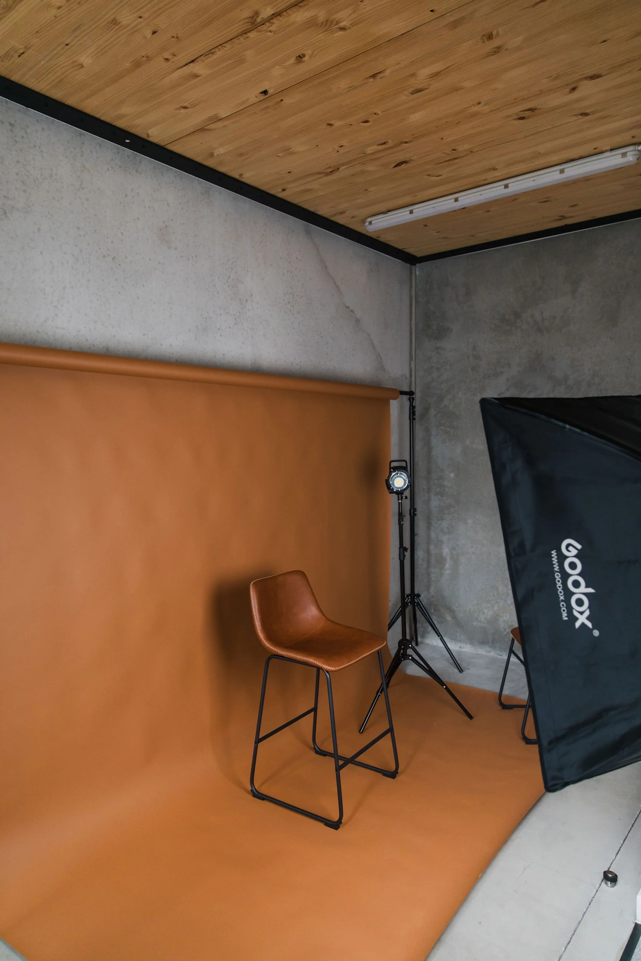 Photo of a photography or video studio setup with an orange backdrop, a brown chair, a black softbox light, and other equipment, with a concrete and wooden wall.