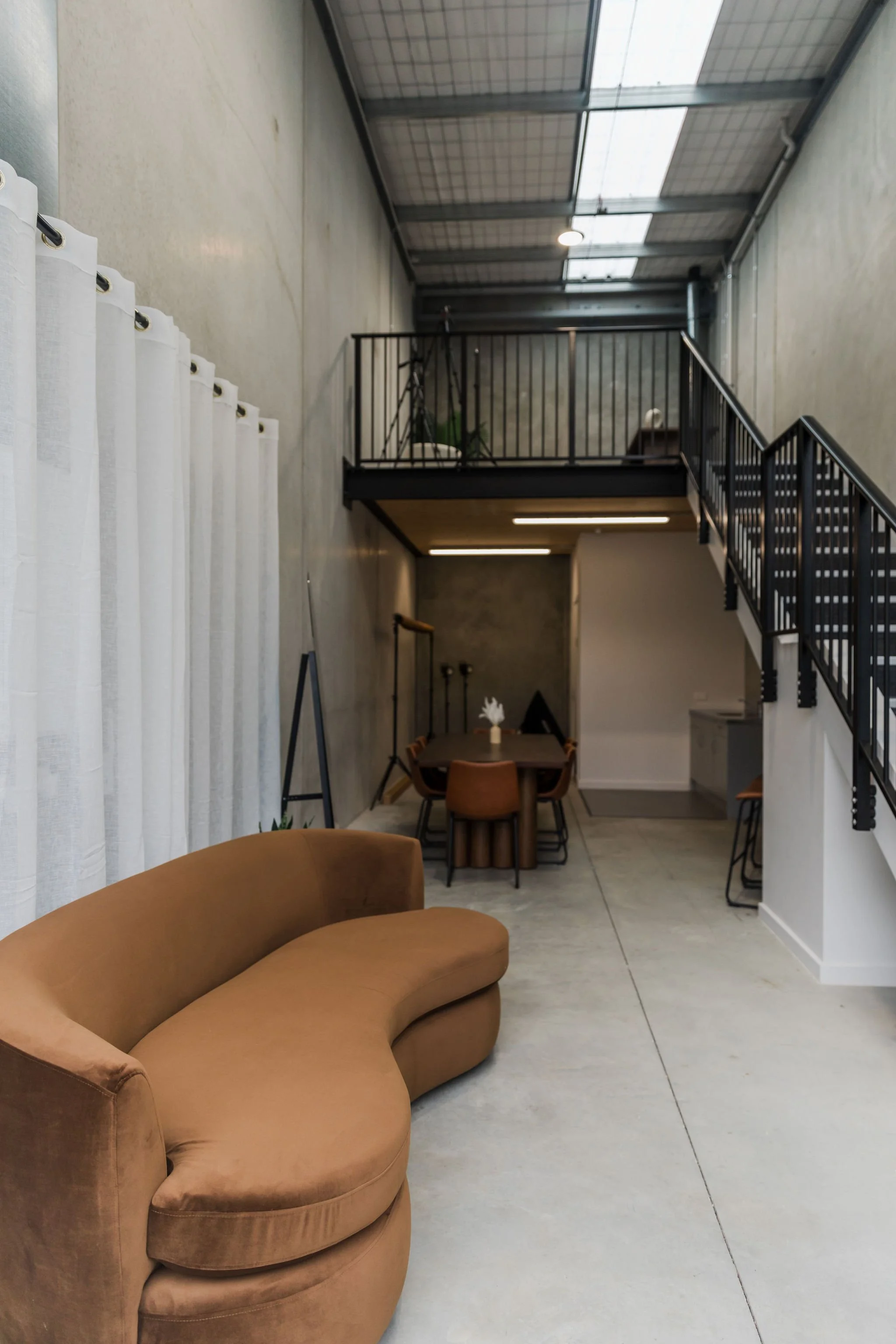 Modern interior space with a curved tan sofa in the foreground, a dining table with four chairs at the back, black metal railing on a loft area, hanging lights, concrete floors, and a ceiling with skylights.