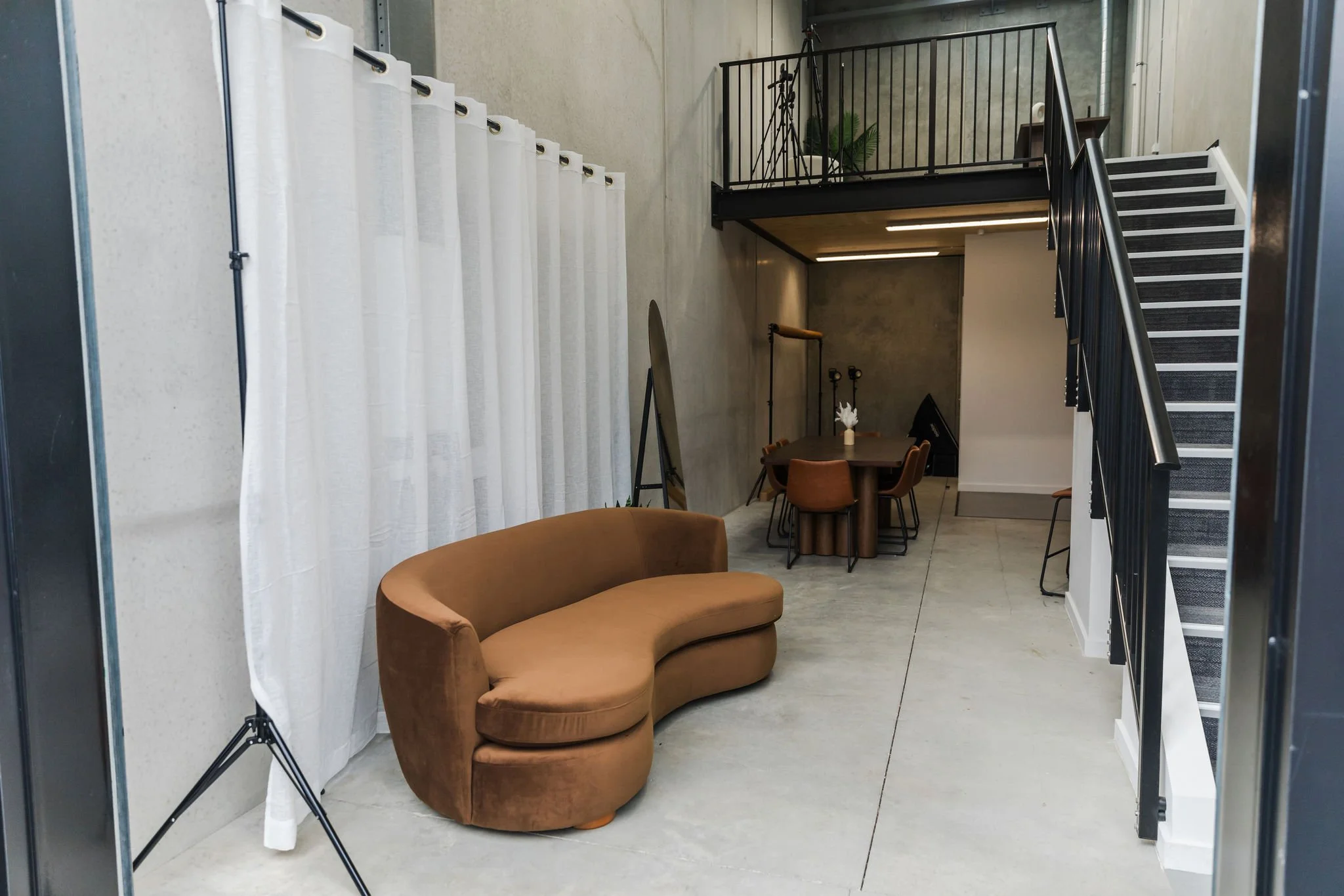 Interior of a modern room with a brown curved sofa, high ceiling, white curtain, staircase with black railing, round dining table with chairs, and studio lighting equipment.