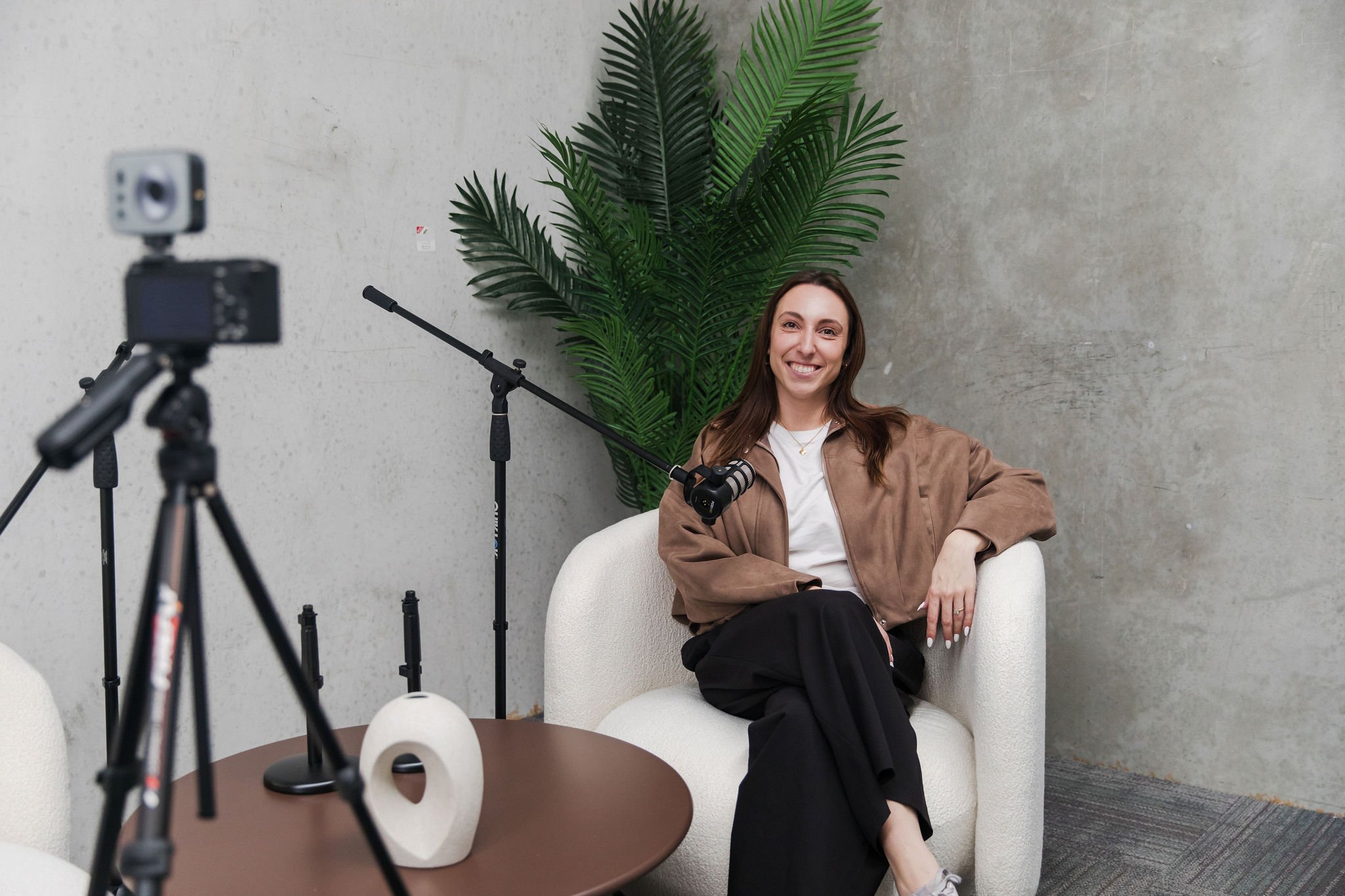 A woman sitting comfortably on a white armchair, smiling, with a large green leafy plant behind her, in a minimalistic indoor setting. A camera on a tripod and a small table with a decorative object are visible in the foreground.
