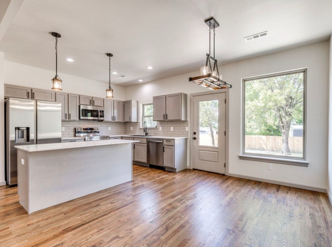 Modern kitchen with light gray cabinets, stainless steel appliances, a white island with a countertop, hardwood floors, and large windows showing trees outside.