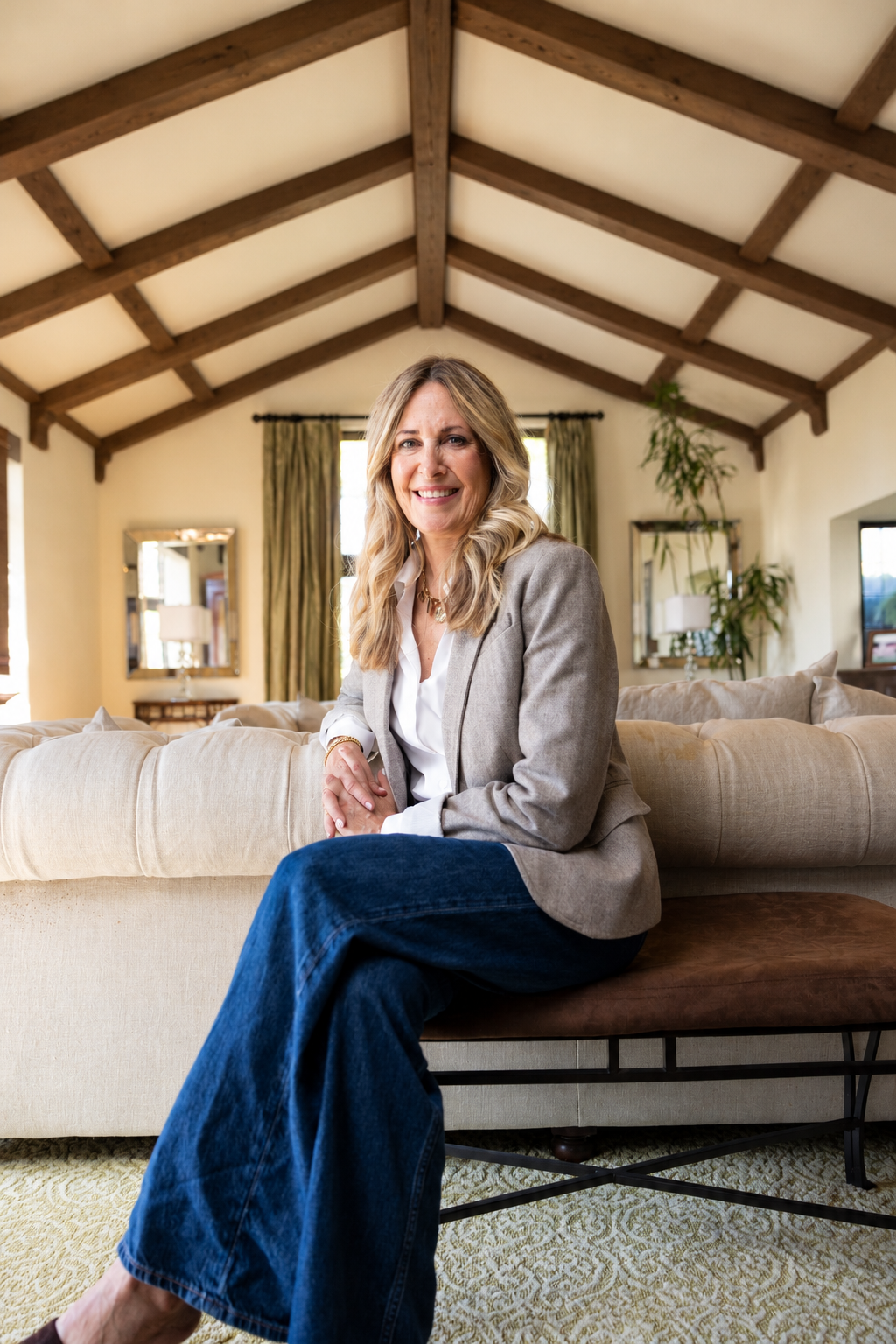 Smiling woman in a gray blazer and wide-leg blue jeans seated on a sofa in a rustic living room with exposed wood beams.