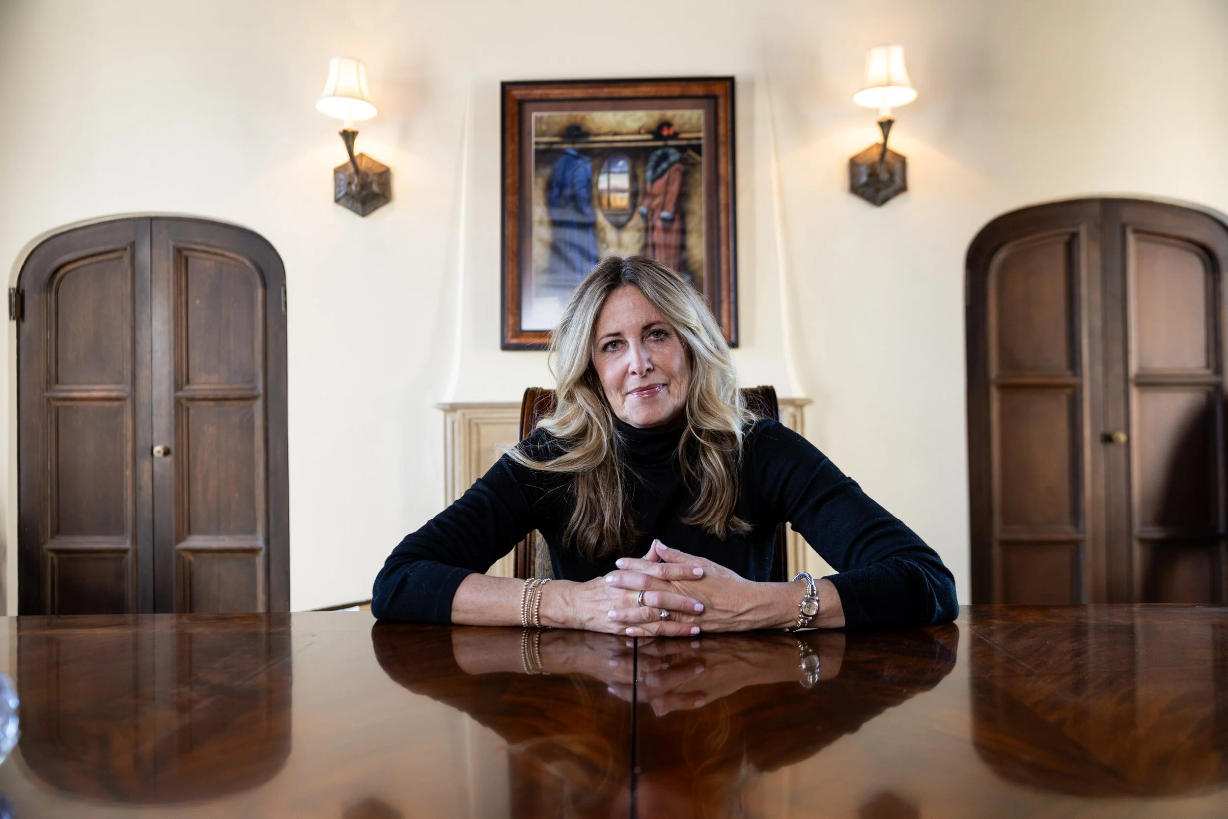 Woman with long blonde hair seated at a polished wooden table in an elegant room