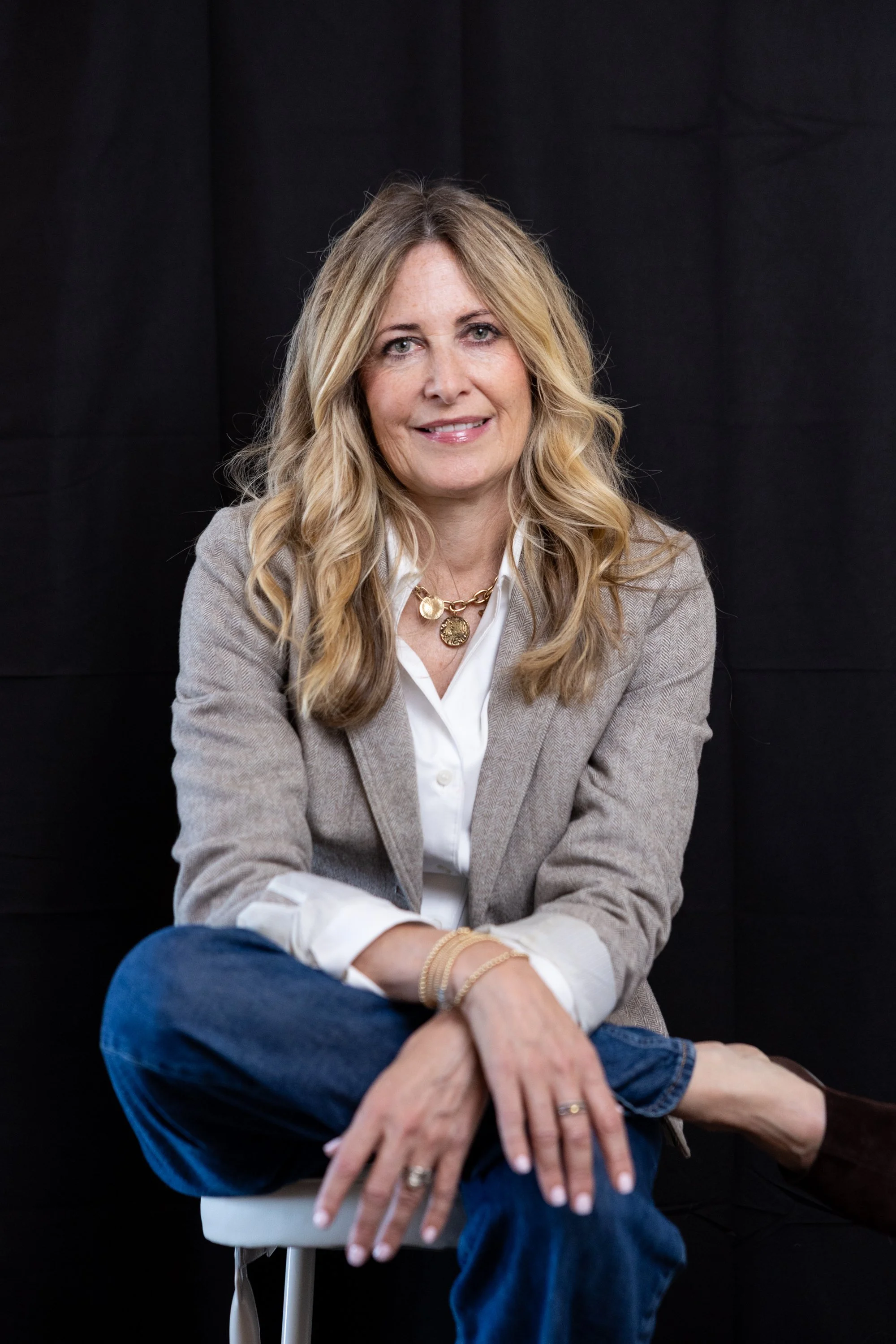 Portrait of a smiling blonde woman in a gray blazer and white blouse seated against a black background.