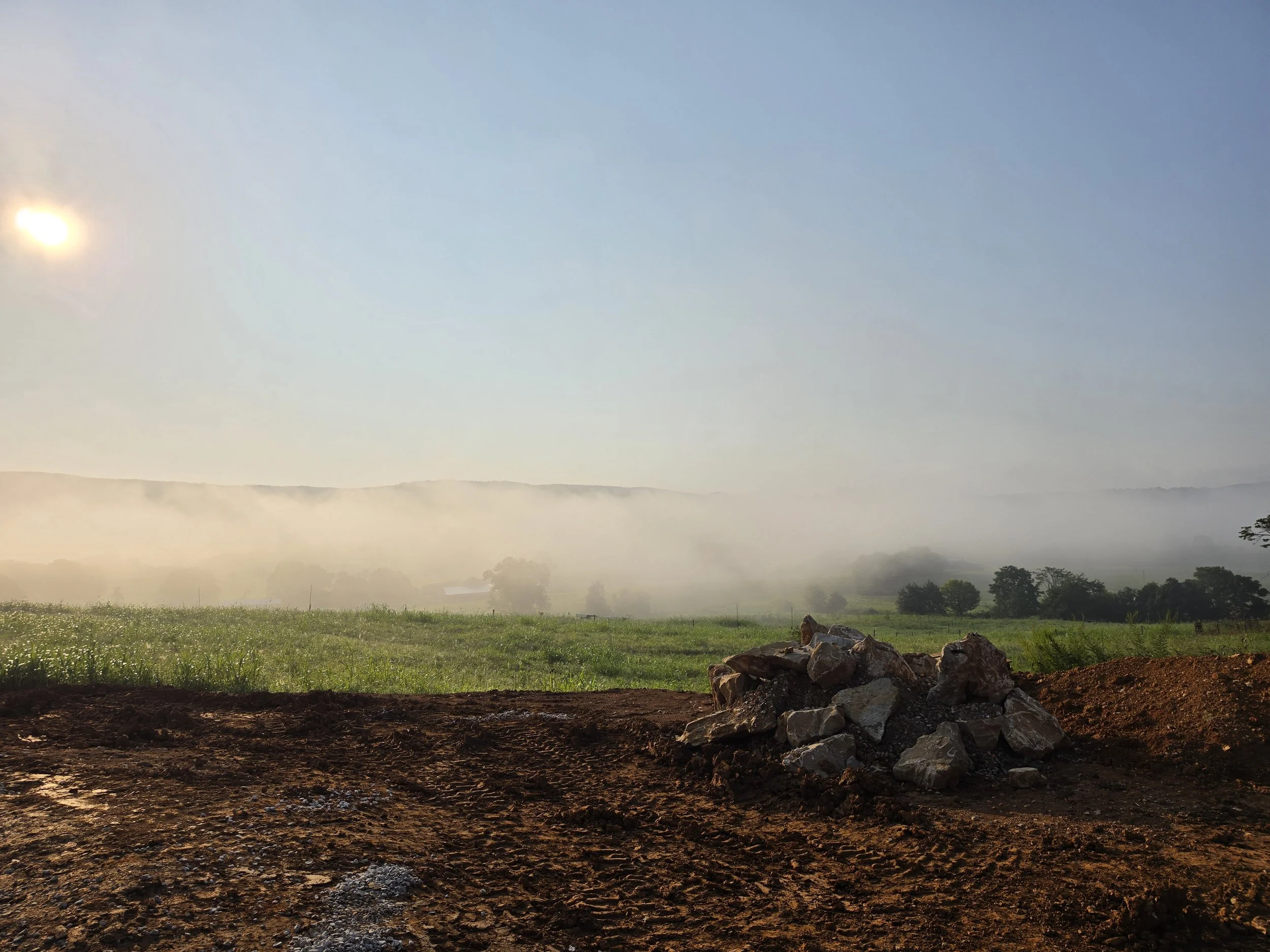 A scenic landscape featuring a dirt foreground with rocks, a lush green field, fog-covered hills in the distance, and a clear blue sky with the sun shining.