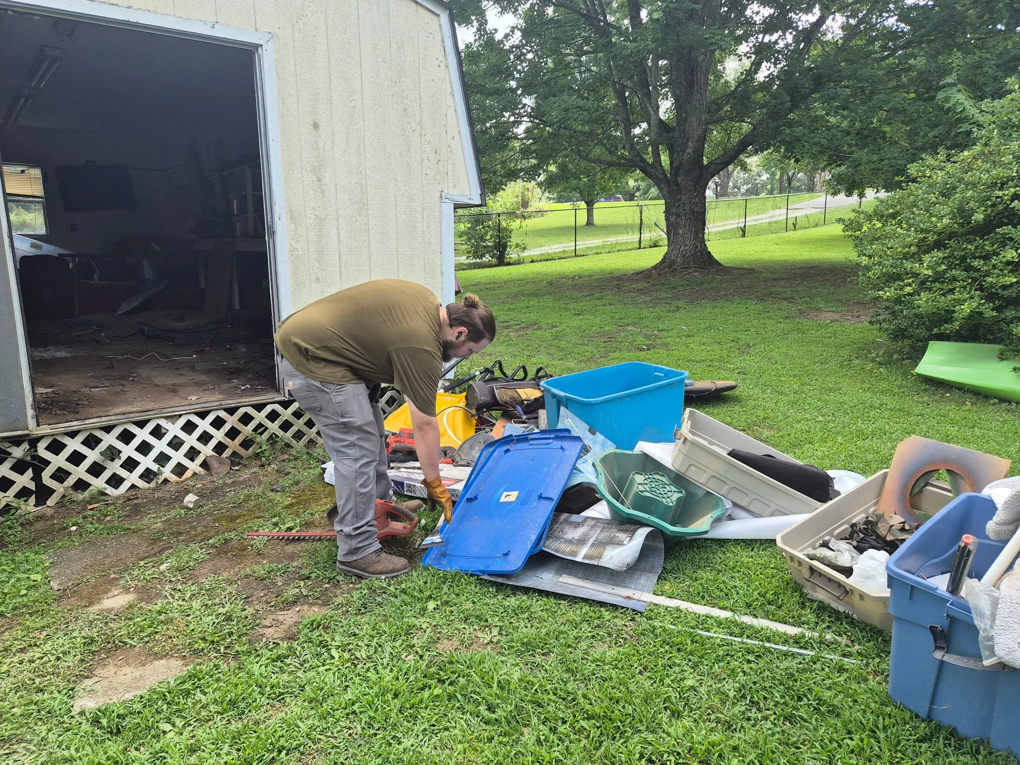 A person bending over near a pile of organized toolboxes and equipment outside a garage, with trees and a grassy yard in the background.