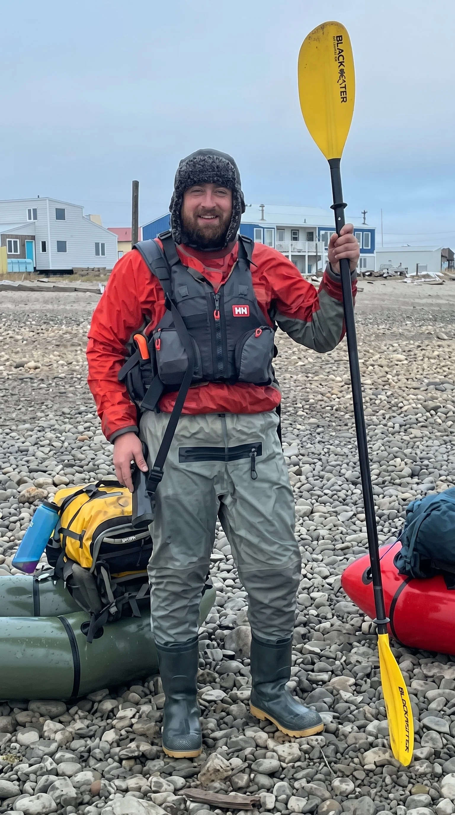 Man in red paddling gear and life vest holding a yellow kayak paddle on a rocky beach beside inflatable kayaks and coastal houses