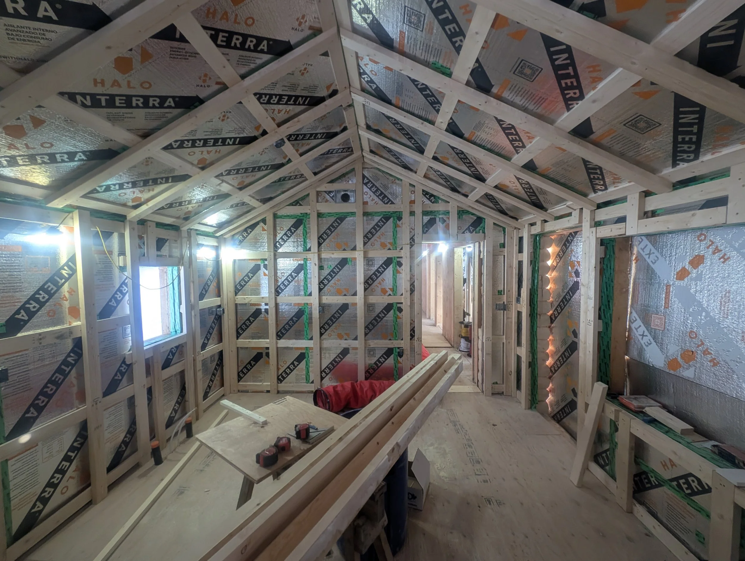 Interior of a house under construction with exposed wood framing, reflective insulation, work lights, lumber, and power tools.