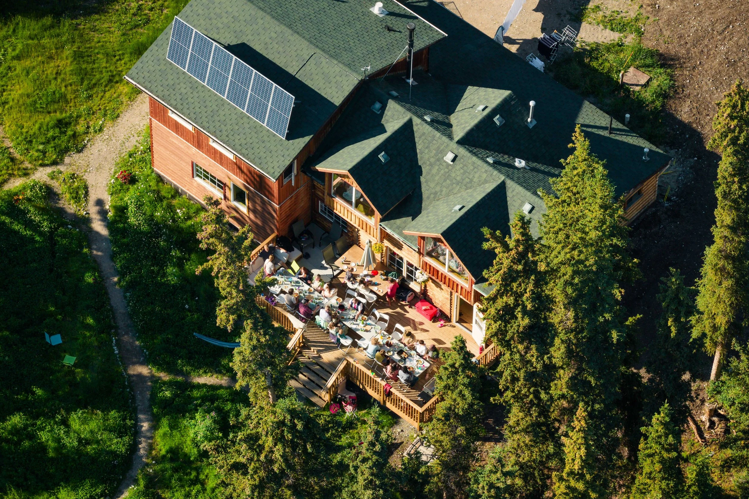Aerial view of a rustic wooden lodge with solar panels, surrounded by trees and gardens, with guests dining on spacious outdoor decks.
