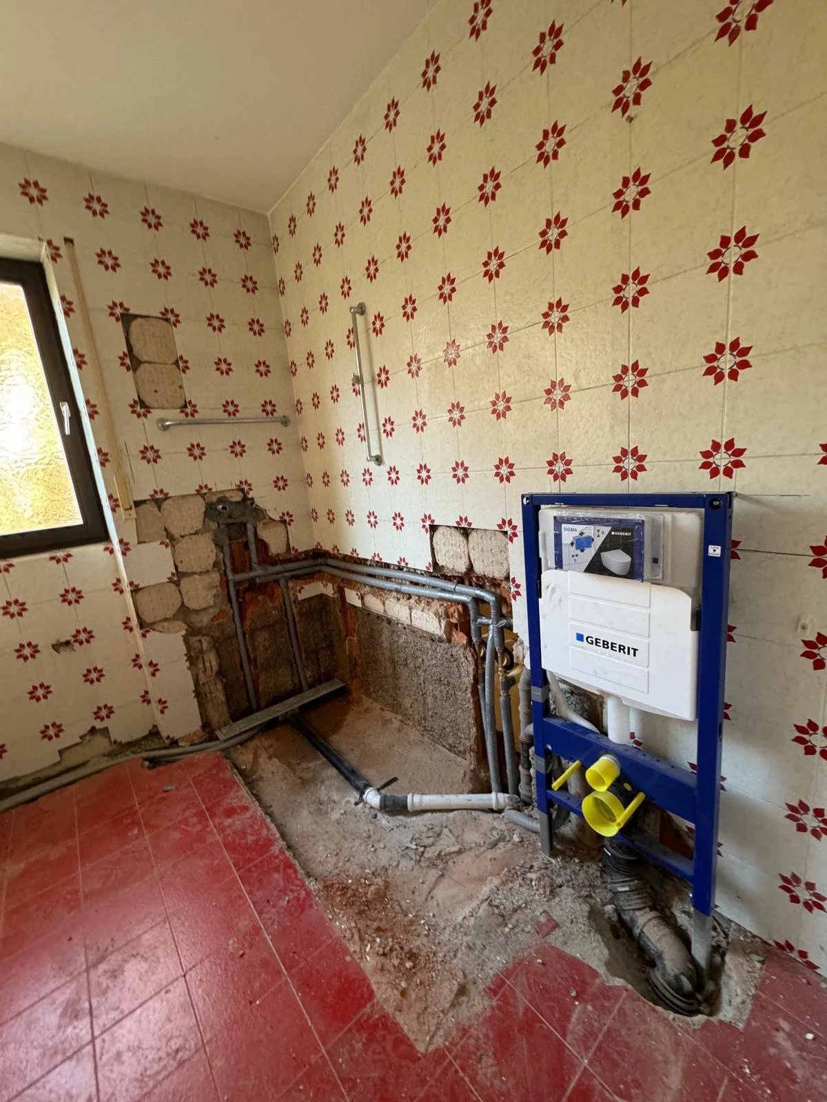 Badezimmer im Umbau mit Rohrleitungen, Wandfliesen mit rotem Blumenmuster, offener Wasserleitungsinstallation und Geberit Spülkasten.