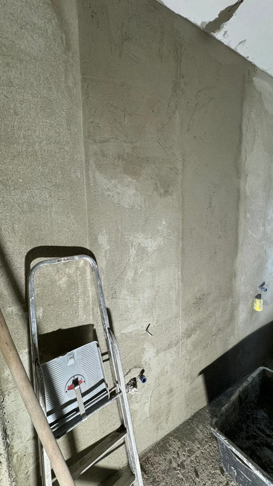 Betonwand in einem Bauprojekt mit Leiter und Mörtelkübel an der Seite, im Hintergrund offene Kabelports.
