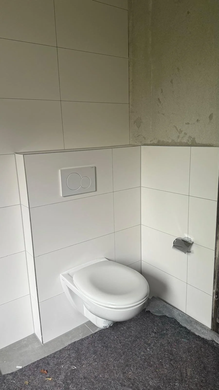 Ein WC mit Wandspülknopf, Wandfliesen und Kabelkanal, unter construction. Der Boden ist unvollständig.