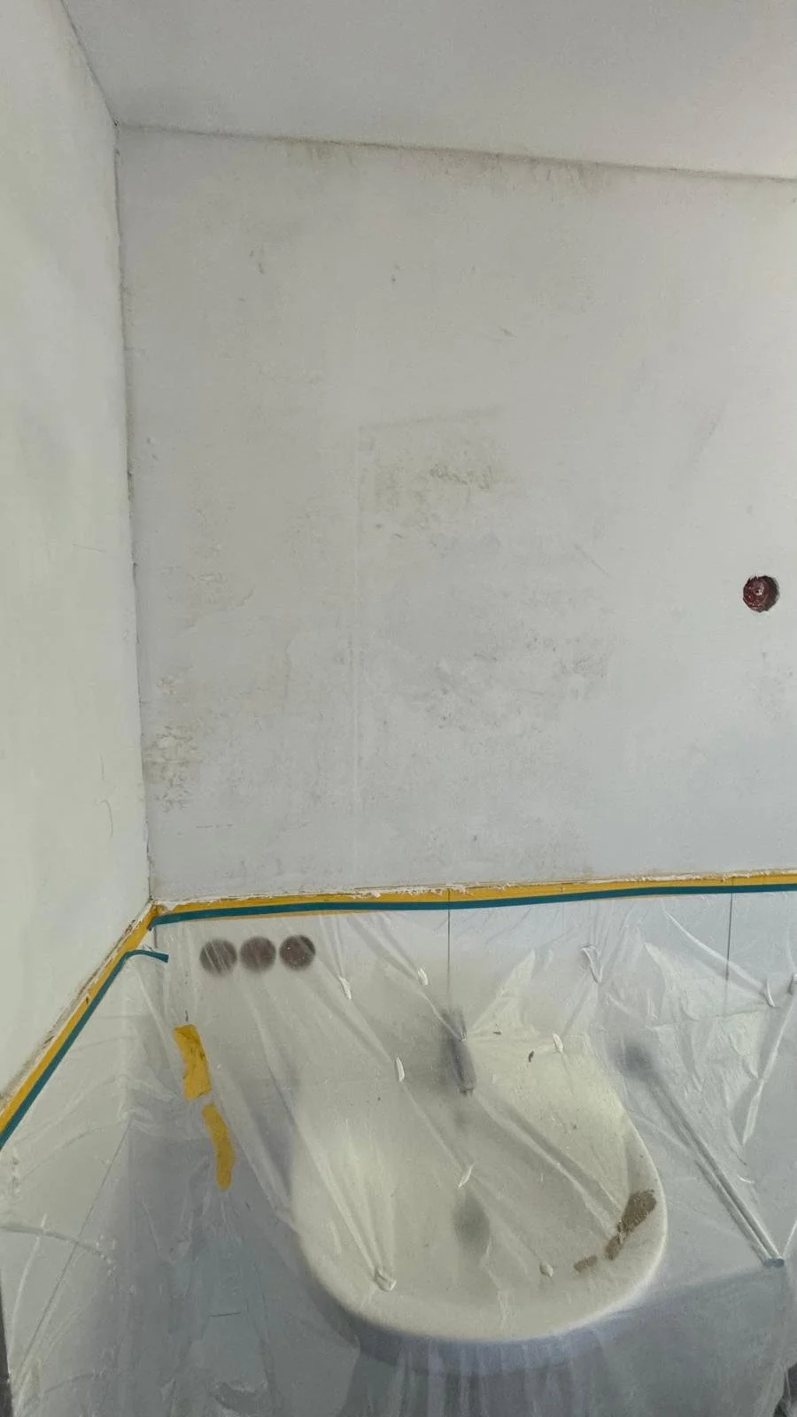 Innenraum eines Raumes unter Renovierung, mit weiß gestrichenen Wänden, Abdeckfolie, und einem Waschbecken. Es sind einige Rohre sichtbar, die auf die Installation von Wasserleitungen hindeuten.