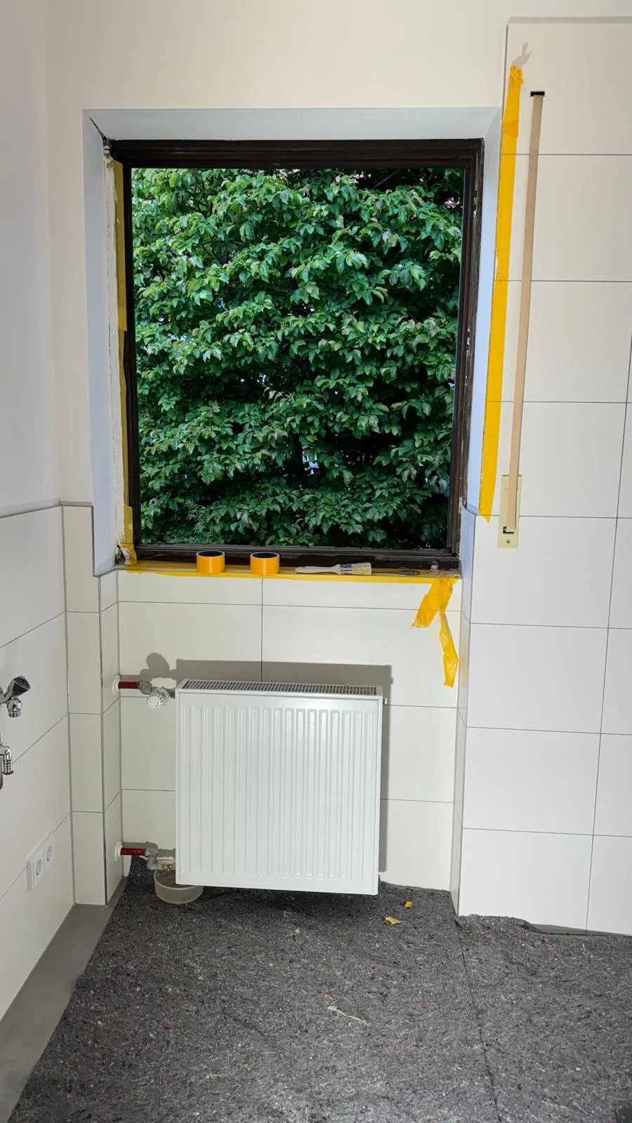 Ein Badezimmer mit einem offenen Fenster, das auf grüne Bäume schaut. Es sind Werkzeuge und Klebeband für die Fenstermontage sichtbar, ein Heizkörper ist unter dem Fenster installiert und die Wand ist teilweise verkleidet.