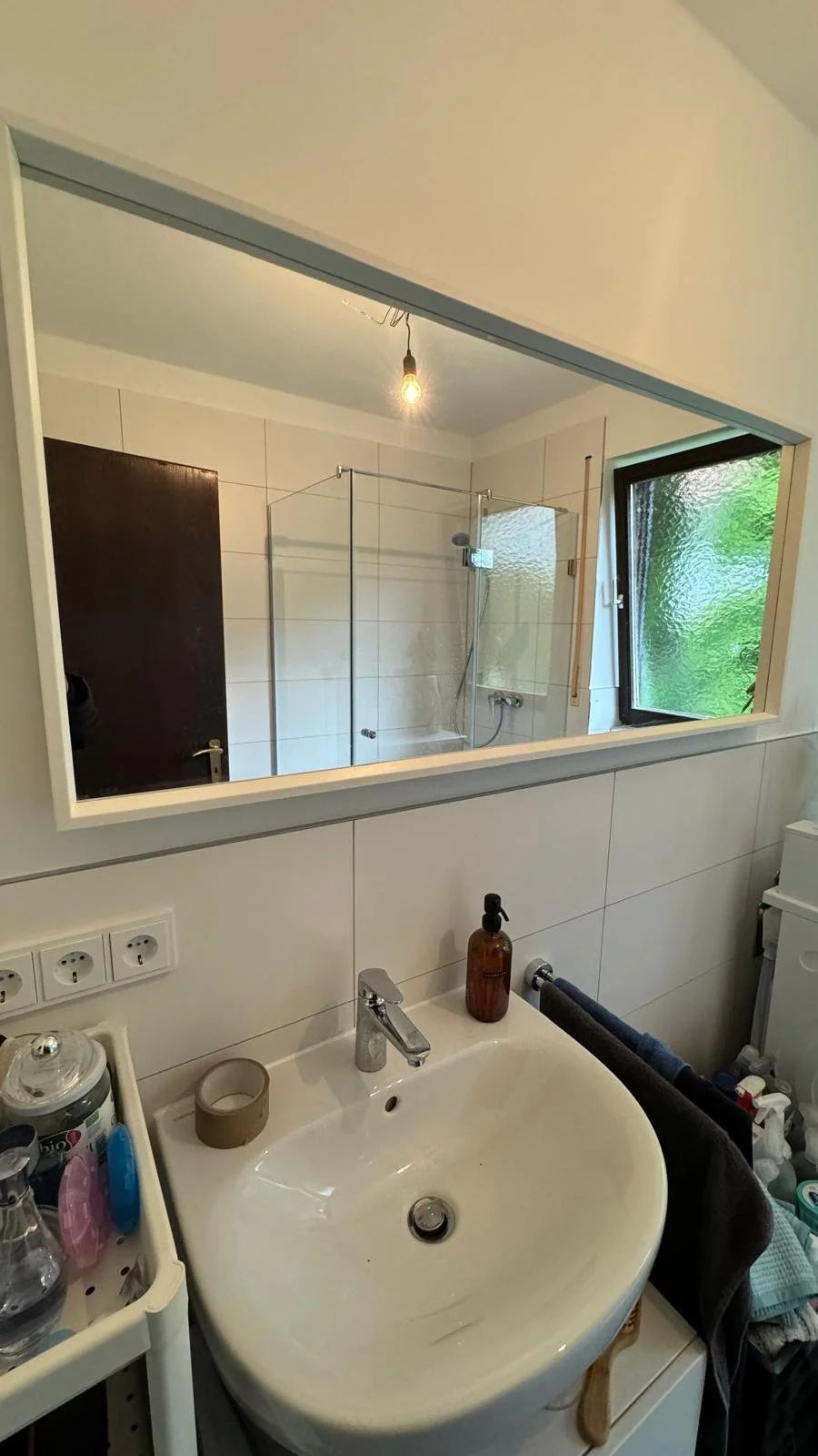 Ein Badezimmer mit Waschbecken, nachdenklich wirkender brauner Seifenspender, Handtücher, Spiegel, Fenster mit Blick nach draußen, Dusche mit Glaswand und Beleuchtung