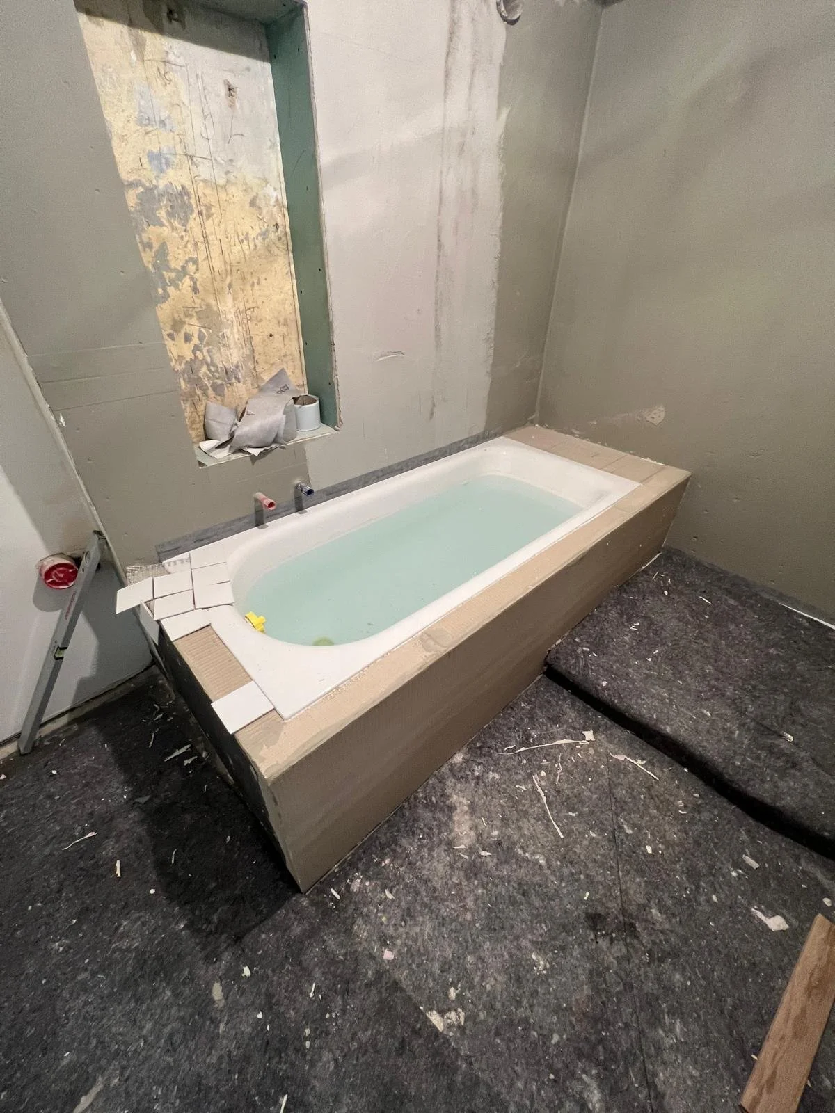 Ein unbeendeter Badezimmerbereich mit einer Einbaubadewanne, die mit Wasser gefüllt ist, auf einer Baustelle oder Renovierungsort. Die Wanne ist in eine Holzkonstruktion eingebettet, mit unverkleideten Wänden und Baumaterialien im Raum.