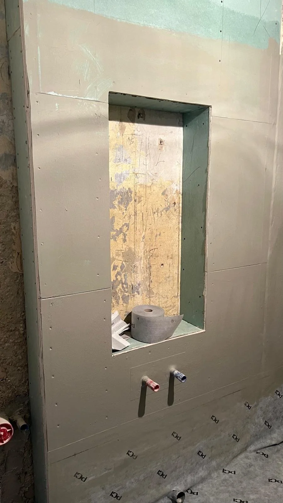 Rohbau einer Wandnische mit Rohrleitungen und Bauschaum, im Hintergrund eine unvollständige Wand mit Farben und Markierungen.