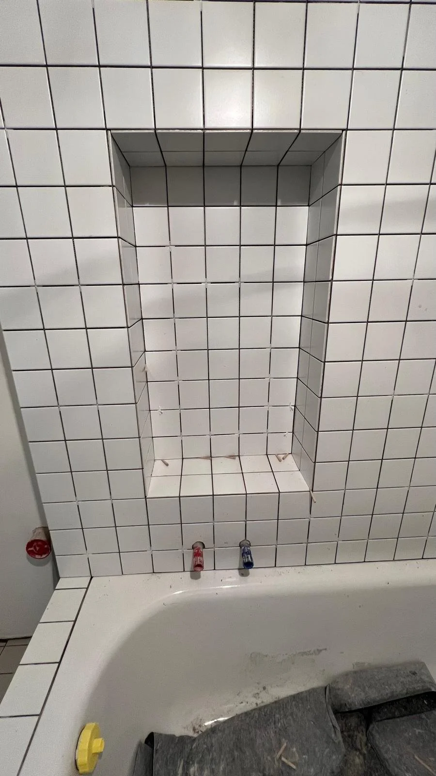 Badezimmer mit weißen Wandfliesen, eine Nische in der Wand, Wasserhähne für Warm- und Kaltwasser, eine Badewanne und eine schwarze Abdeckung auf dem Boden.