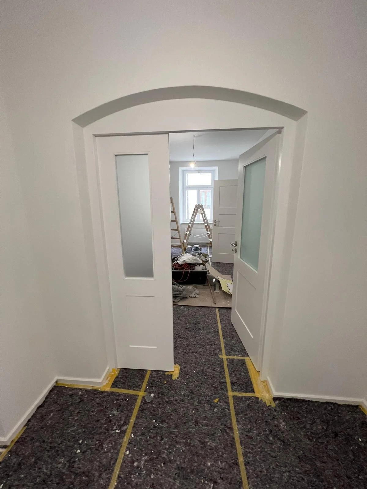 Innenraum einer renovierten Wohnung, Blick durch offene doppelte Türen in ein weiteres Zimmer mit Treppestufen, Fenster und Bauarbeiten, gelbe Markierungen auf dem Boden, Werkzeuge und Materialien auf dem Boden.