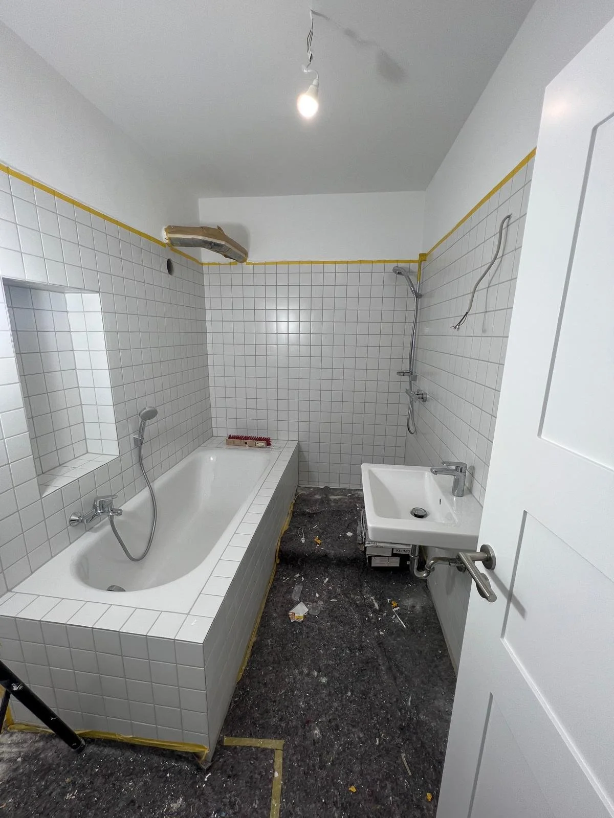 Badezimmer im Bau mit weißer Wand- und Fliesenverkleidung, Badewanne, Waschbecken, unfertiger Deckeninstallation und dunklem Boden.