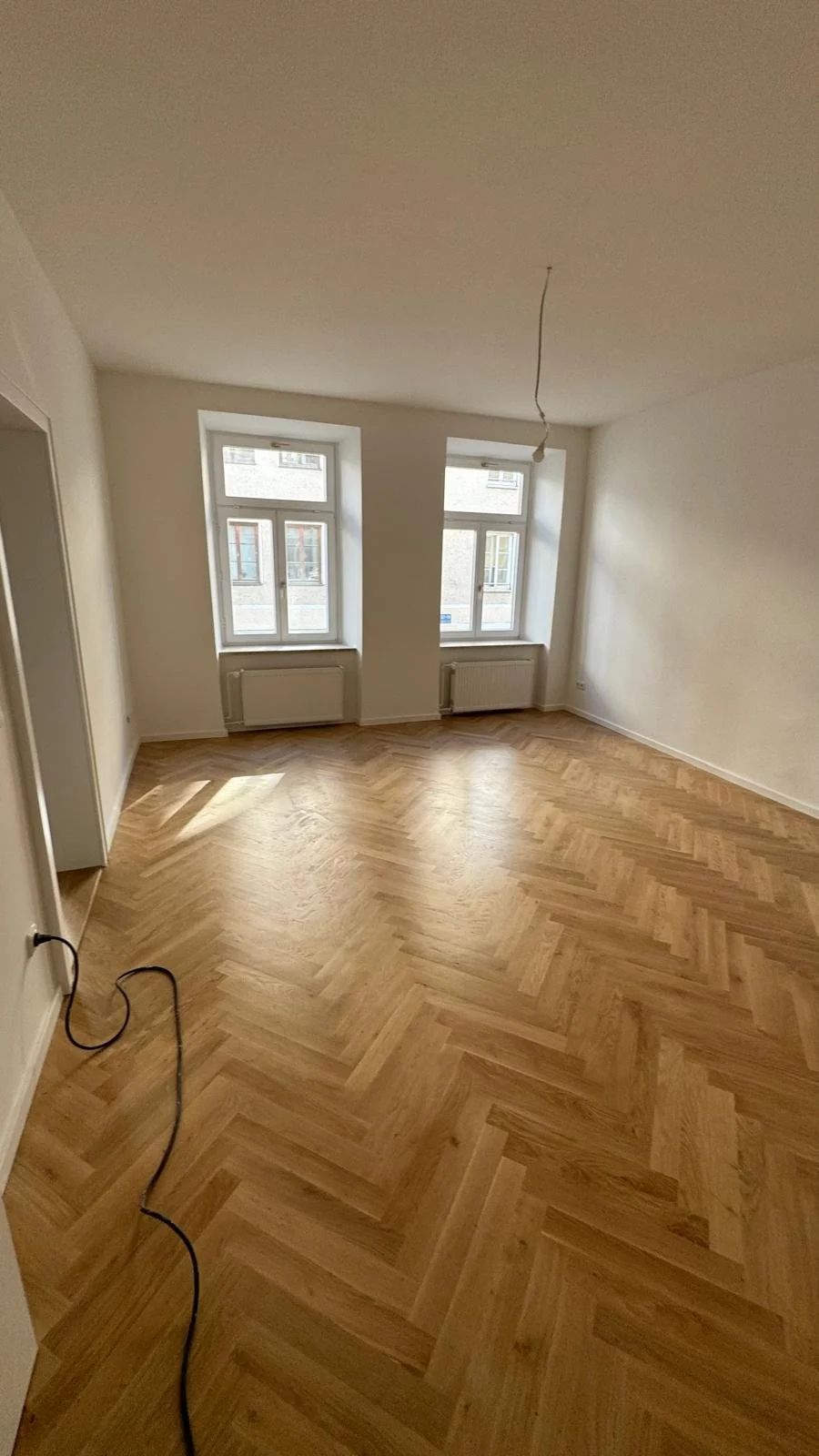 Leeres, frisch renoviertes Zimmer mit Holzboden, zwei große Fenster und eine Deckenlampe, Kabel auf dem Boden.