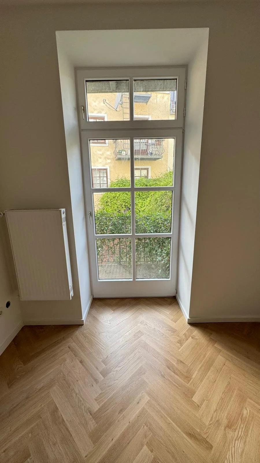 Innenraum mit Holzfußboden, weißer Wand, französischem Balkon mit Glasfenster, Blick auf grüne Bäume und ein Nachbargebäude.
