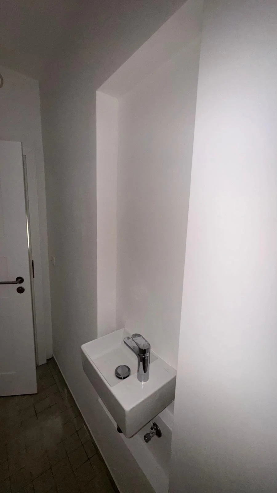 Kleiner Waschtisch mit Wasserhahn in einem kleinen Badezimmer, weiße Wände und braune Fliesen auf dem Boden.