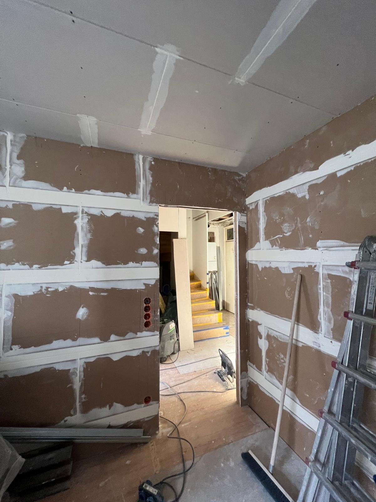 Innenraum eines Hauses, Baustelle mit unvollendeten Wänden aus Gipskarton, Bauwerkzeuge, Leiter, Treppe im Hintergrund, Elektrik-Installation in Arbeit.