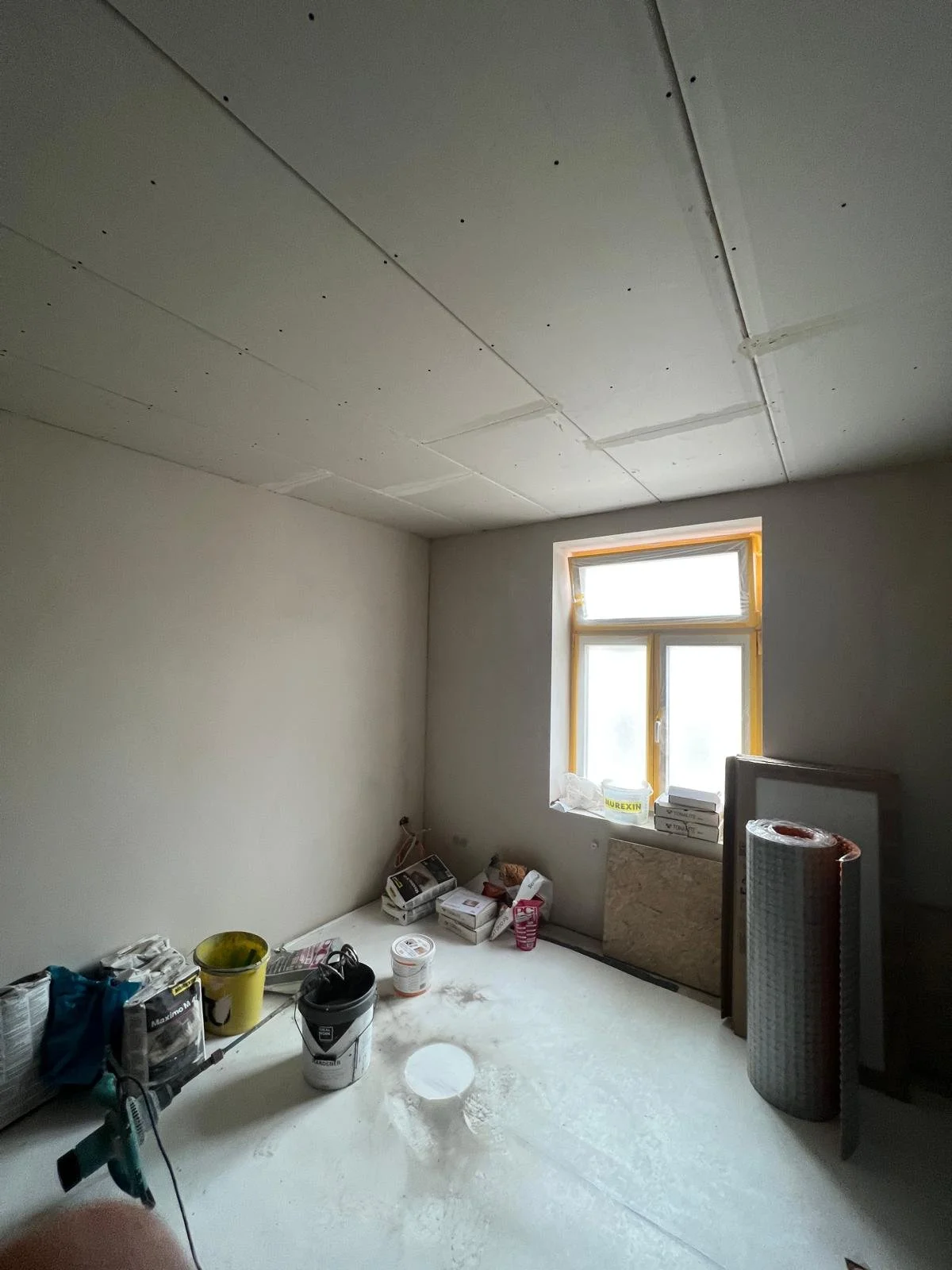 Innenaufnahme eines unvollendeten Zimmers mit teilweise fertiggestellter Decke und Baumaterialien auf dem Boden, darunter Eimer, Puder, Werkzeuge und Verpackungen, sowie eine Fensterfront mit natürlichem Licht.