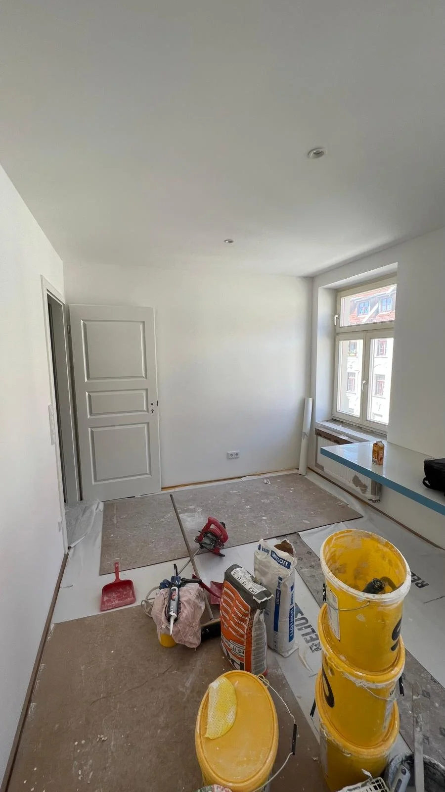 Ein unmöbliertes Zimmer wird renoviert, mit Malerutensilien, Farbeimer, Pinsel, Spachtel und Werkzeug auf dem Boden.