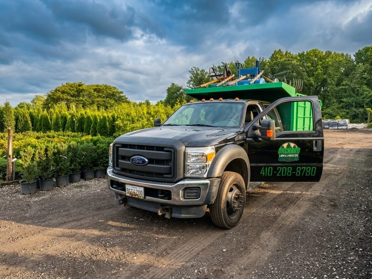 A black Ford pickup truck parked outside on a dirt lot, with gardening tools and equipment on top, and a green and black sign on the door reading 'Orchard Lawn & Landscape' with a phone number. There are trees and neatly arranged potted plants in the background under a cloudy sky.