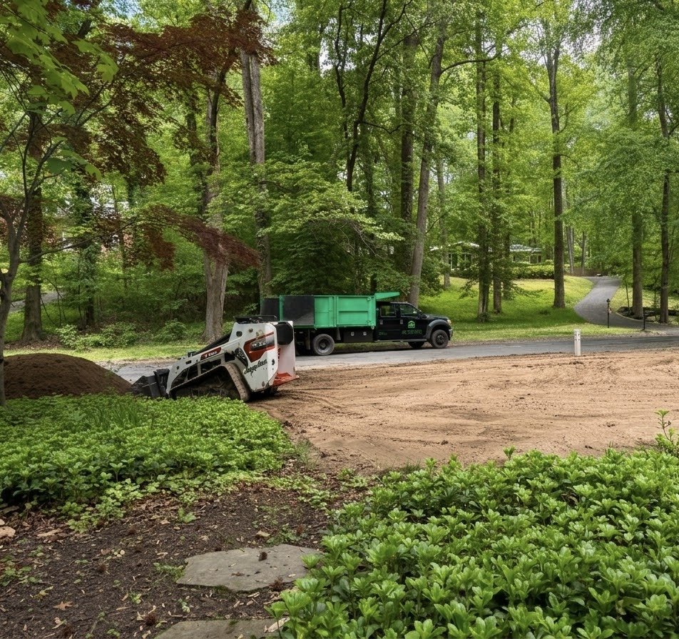 A construction site surrounded by trees with a skid-steer loader and a dump truck parked on a dirt area, and a winding road in the background.