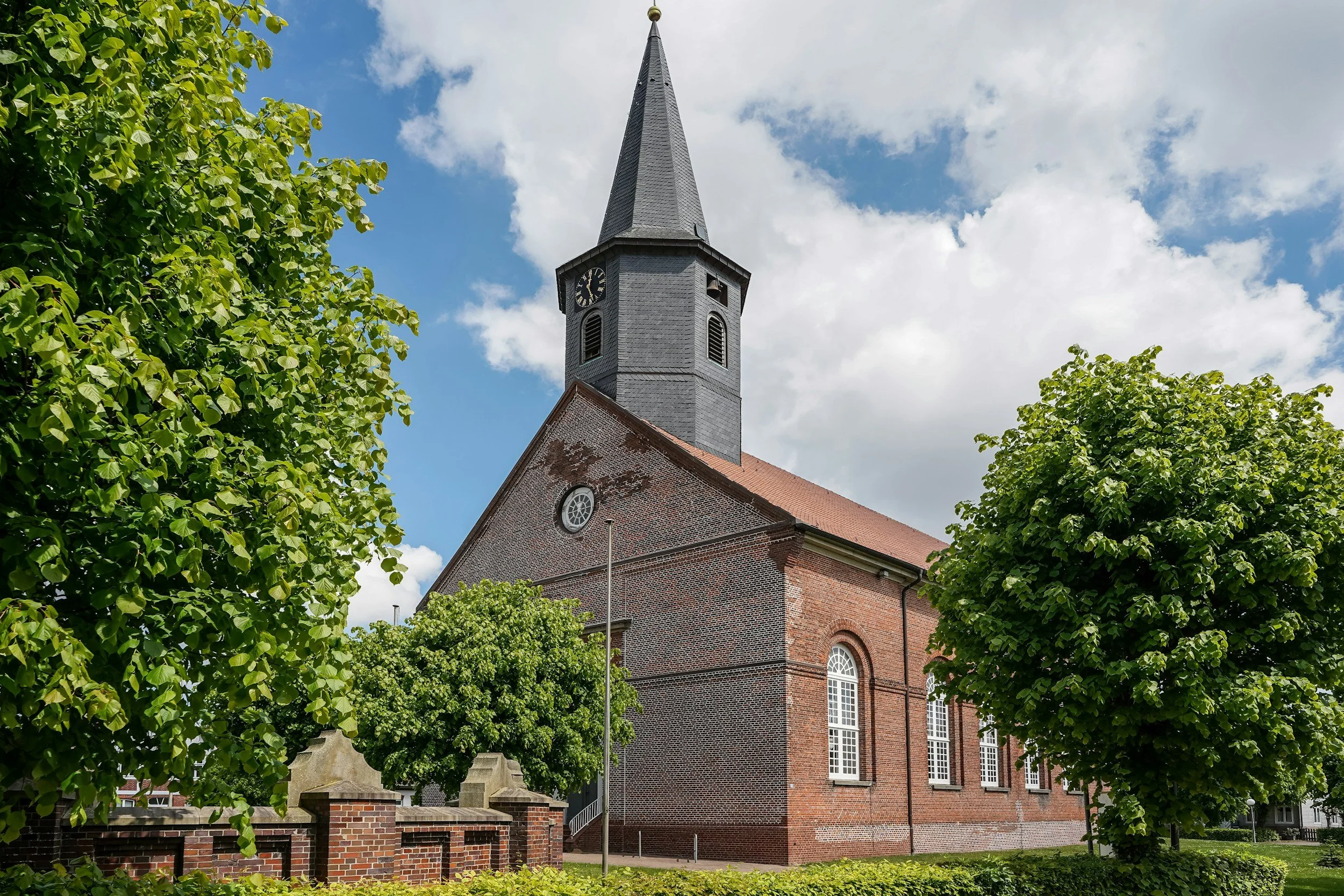 A brick church with a tall steeple and clock, surrounded by green trees and a partly cloudy sky.
