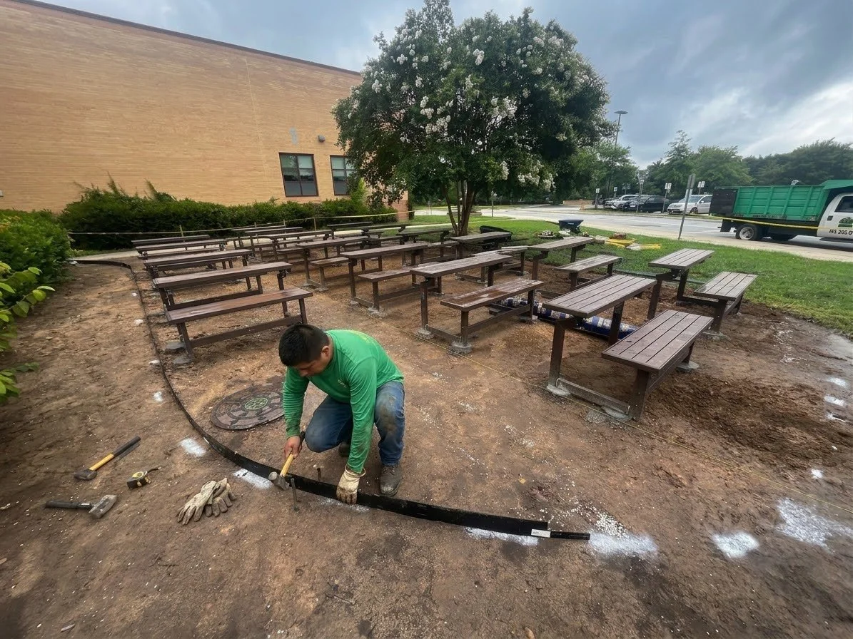 A worker installing outdoor benches under a flowering tree in a park or public space, with construction tools and gloves nearby, and a building and street in the background.