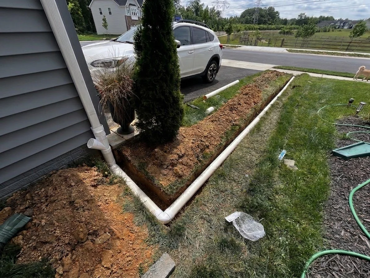 A garden bed with freshly dug earth, white PVC pipes for irrigation, and a small tree next to a house. There's a parked car on the street, a grassy area, and a sidewalk in the background.