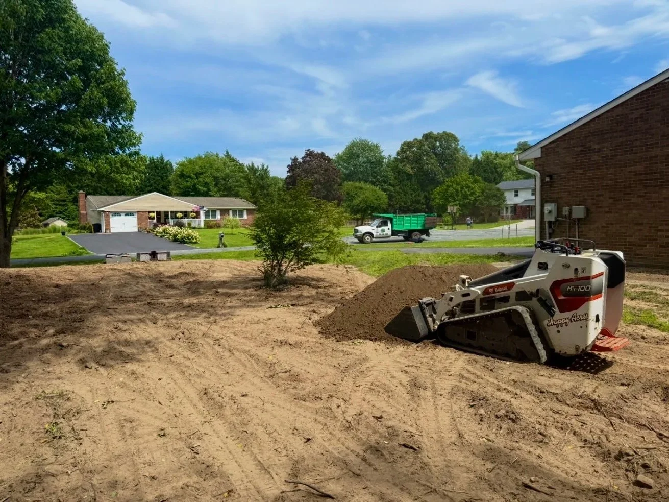 A robotic land registrar machine on dirt in a backyard, with a small bush in the center, surrounded by houses, trees, and street in a suburban neighborhood.