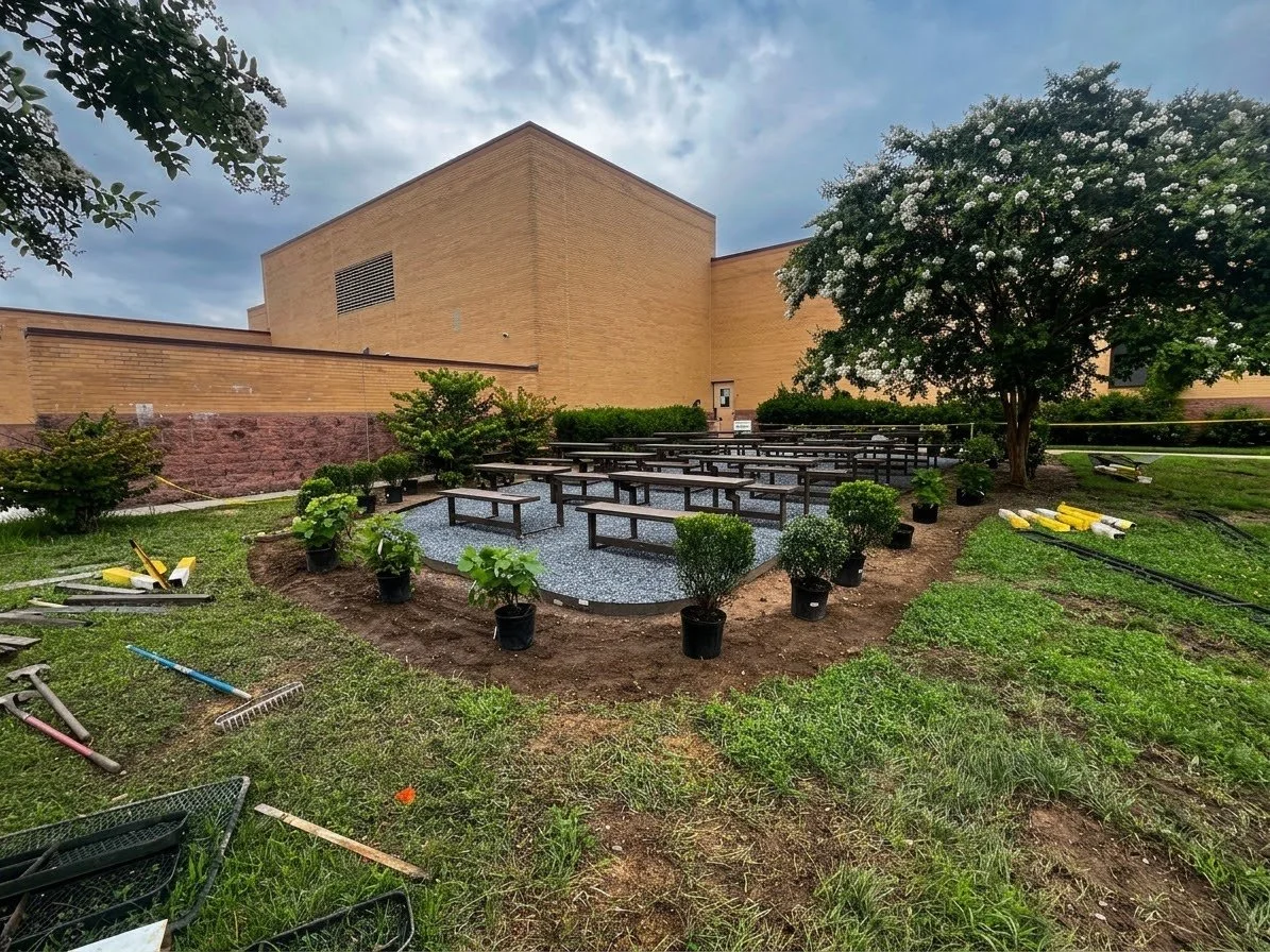 Outdoor patio with arranged potted plants, bushes, and a flowering tree, adjacent to a large building with a brown brick exterior, under a cloudy sky.