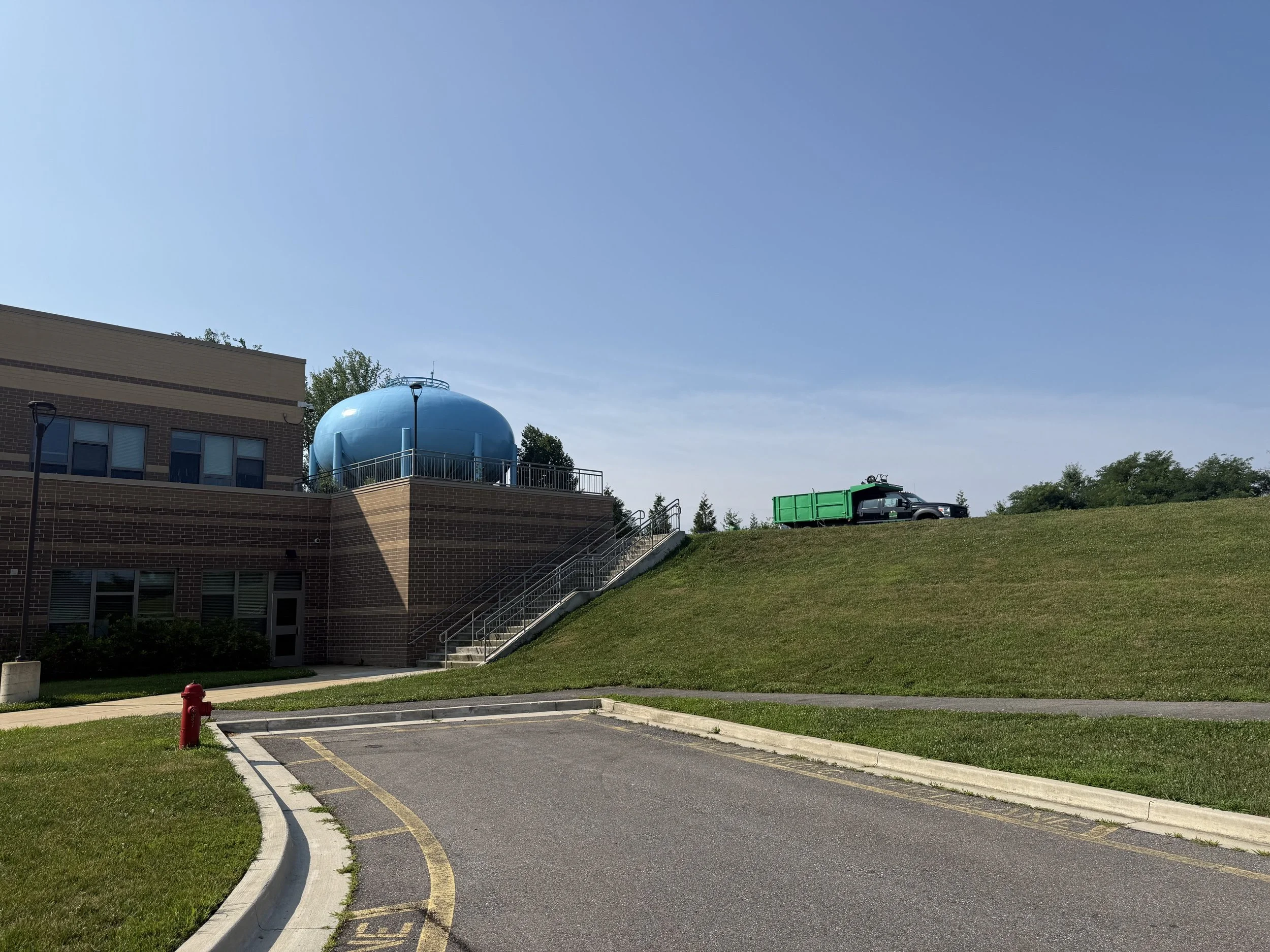 Empty parking lot with a fire hydrant, a grassy area, and a building with a blue water tower on a hill in the background under a clear blue sky.