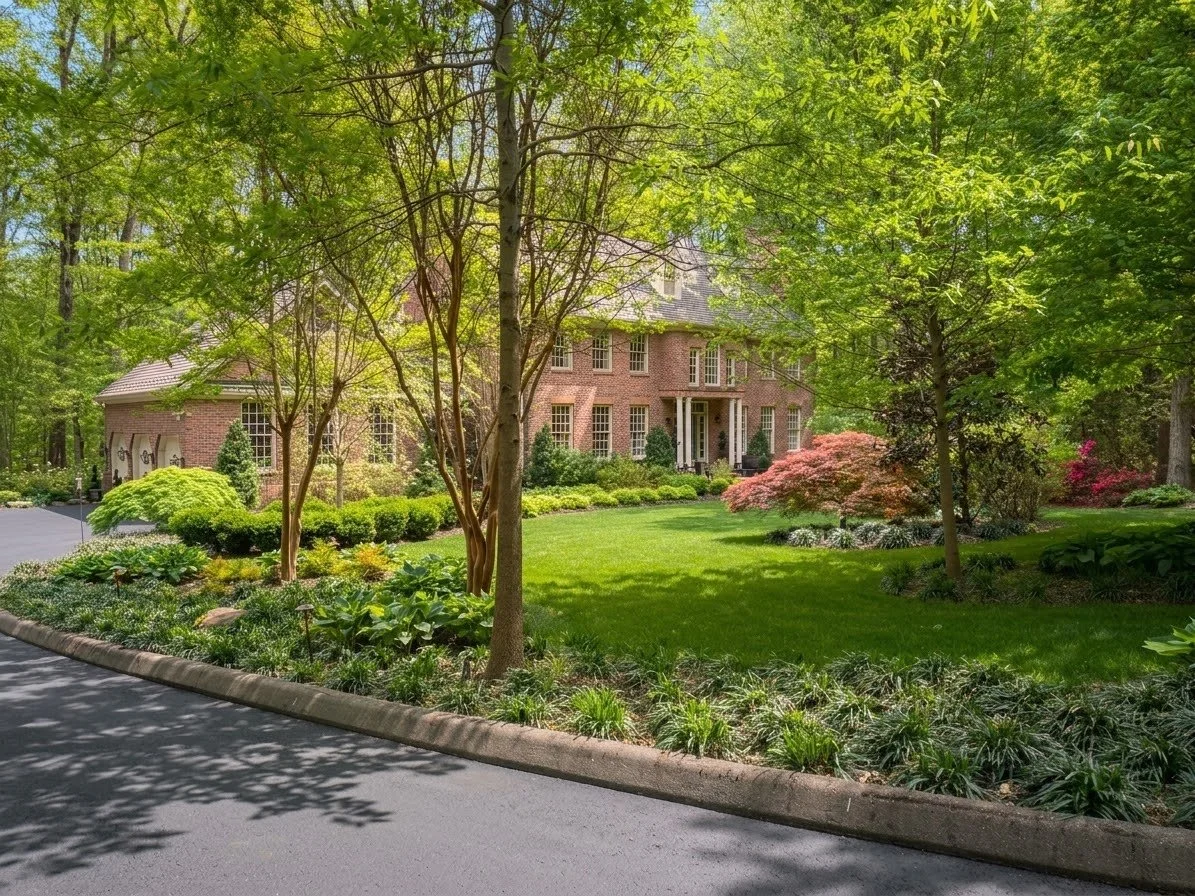 Large brick house surrounded by lush green trees and well-maintained garden with colorful shrubs and flowering plants, with a driveway in the foreground.